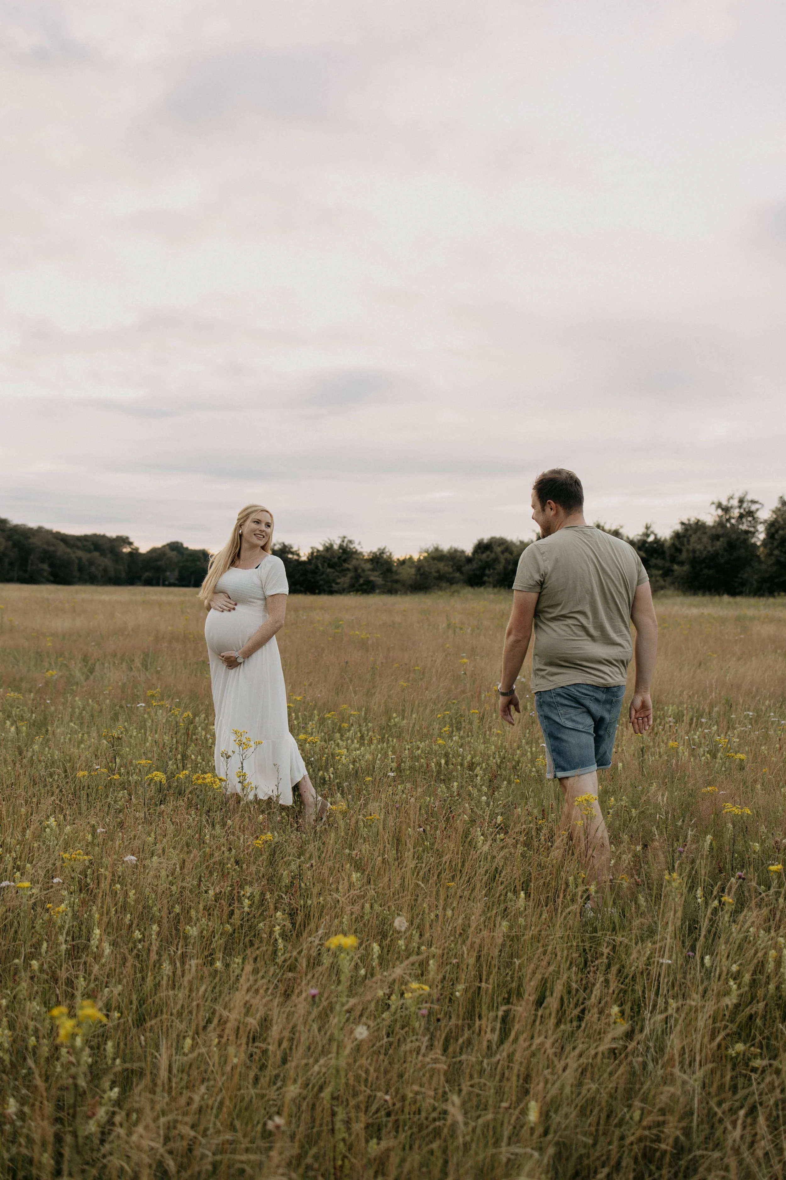 A pregnant woman in a white dress standing in a grassy field, smiling and looking at a man in casual attire who is walking towards her.