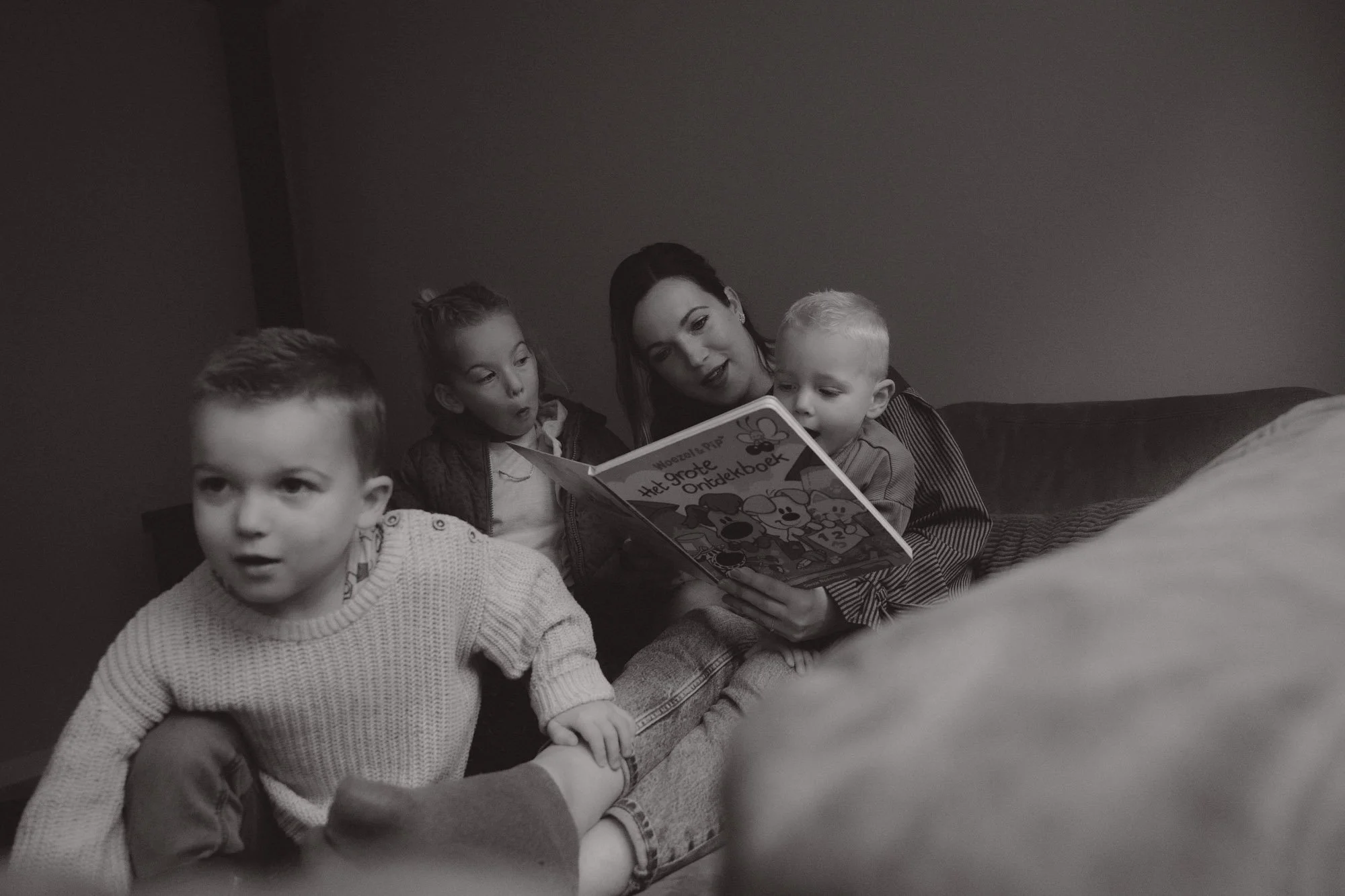 A woman reading a cartoon book to three children sitting on a couch in a dimly lit room.