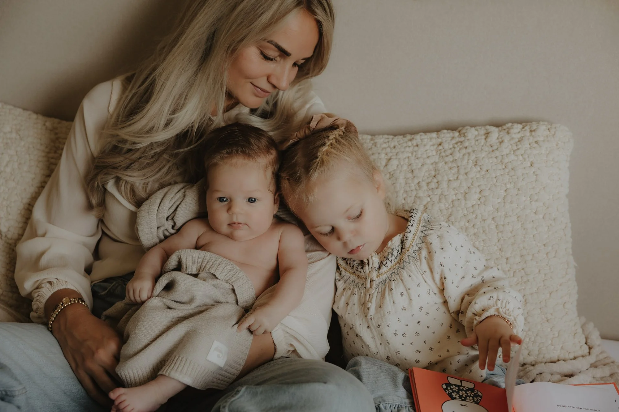 A woman with long blonde hair sits on a couch with two young children, a girl and a baby, all engaged in looking at a book together. The woman is smiling, and the children are focused on the book, which the girl is flipping through.