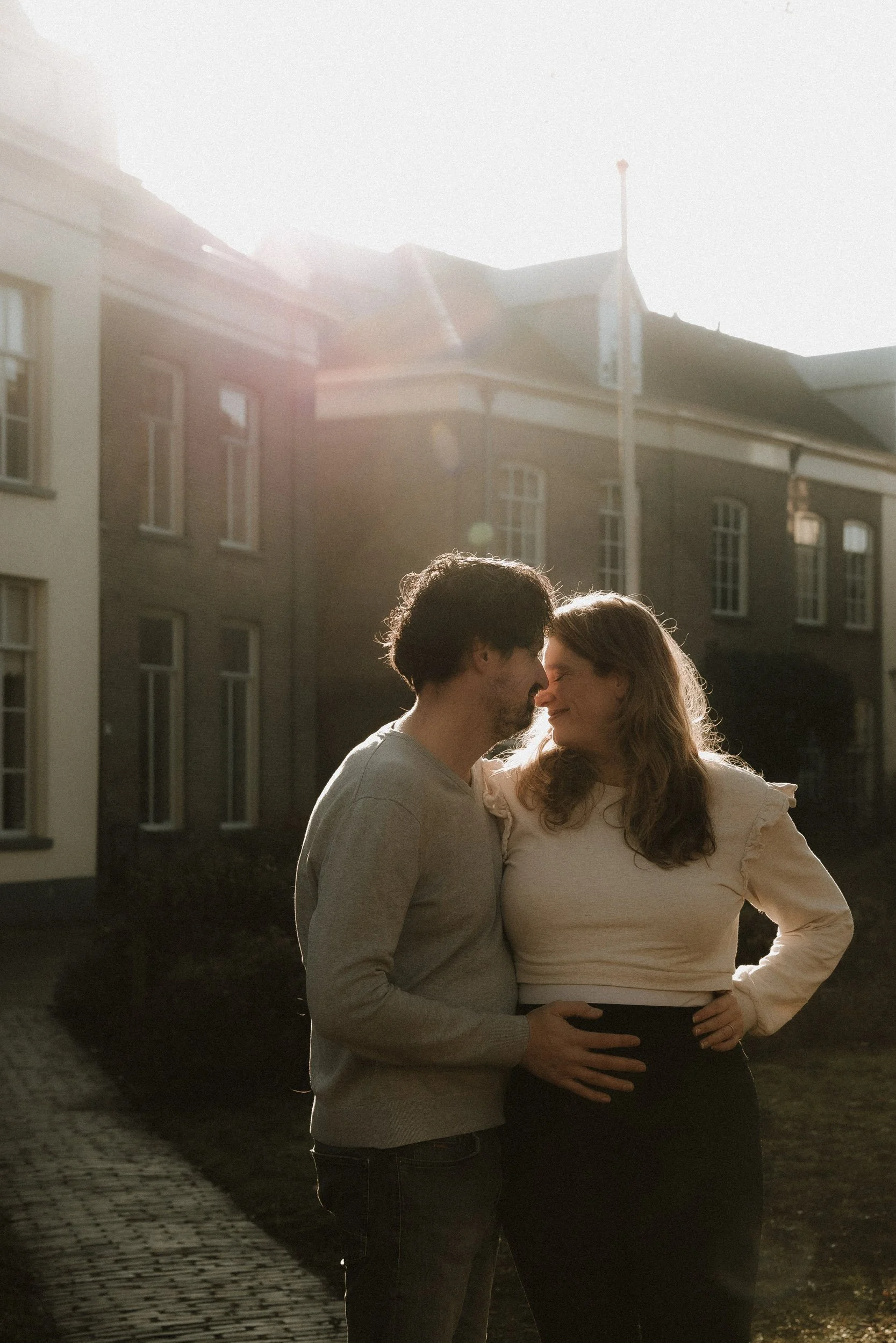 A couple is standing close together outside during sunset, touching foreheads and smiling, with residential buildings in the background.