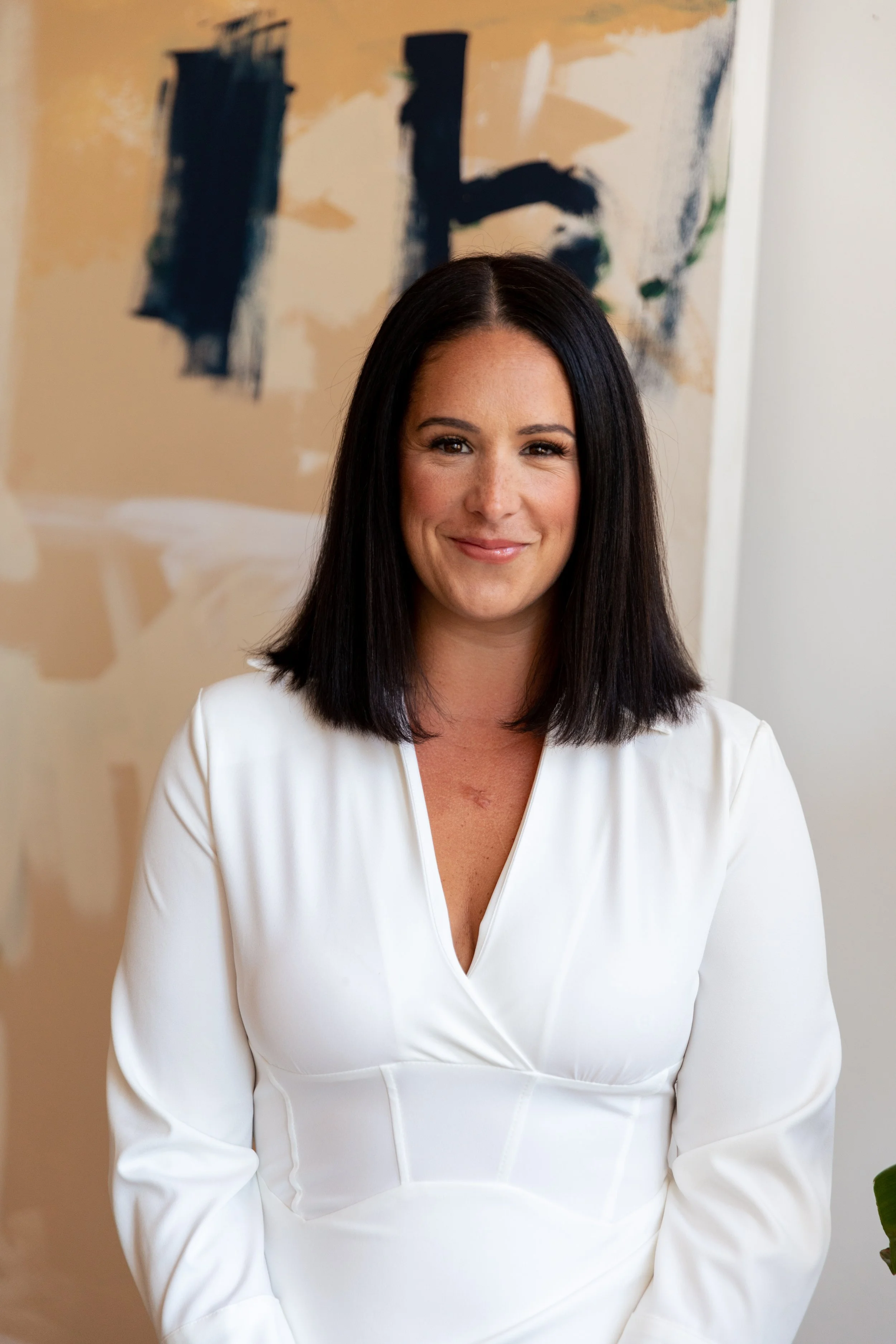 A woman with shoulder-length dark hair, wearing a white dress, smiling in front of an abstract painting with beige, black, green, and blue colors.