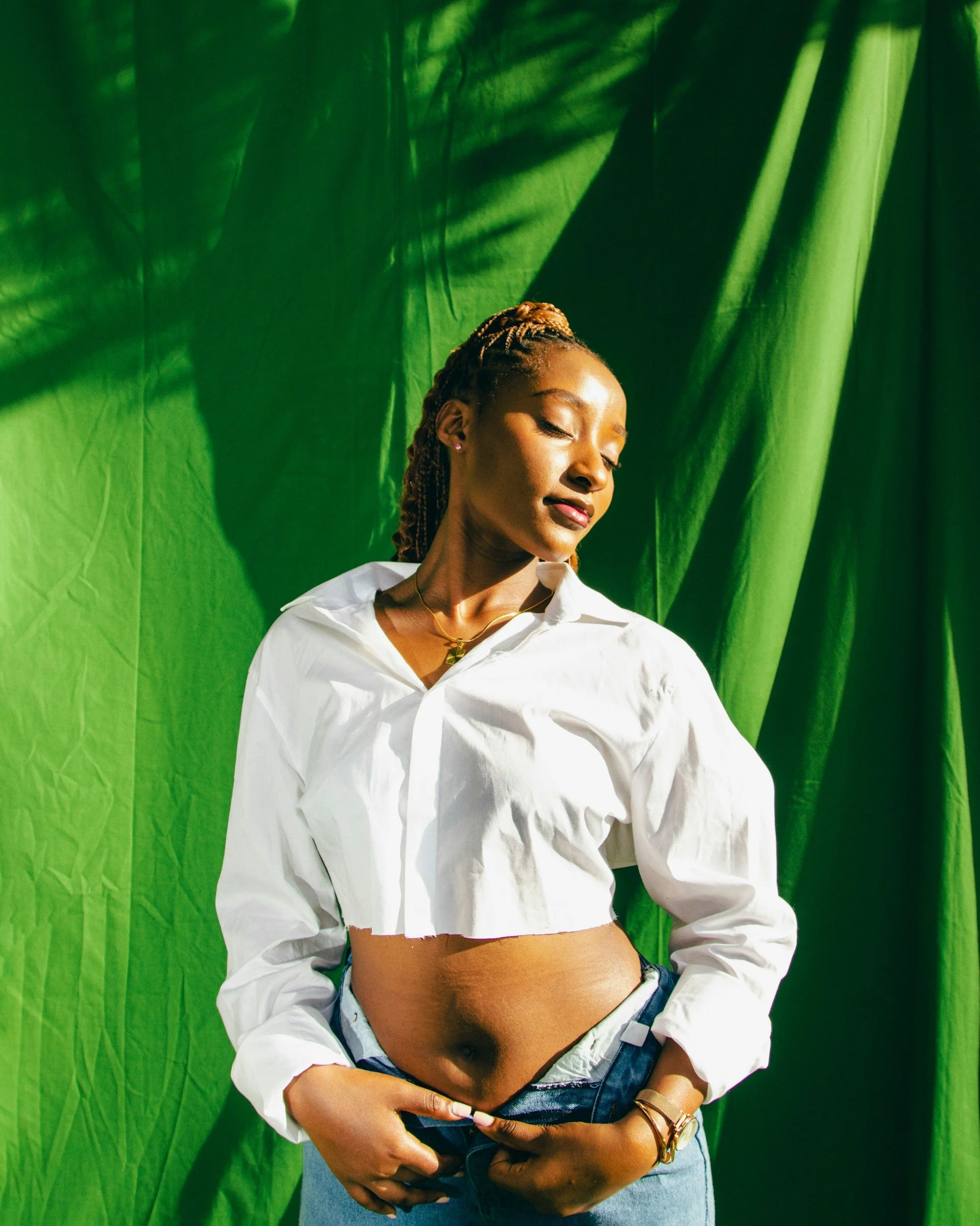 A woman standing in front of a green background, wearing a white cropped blouse and blue jeans, with her eyes closed and head tilted slightly to the side.