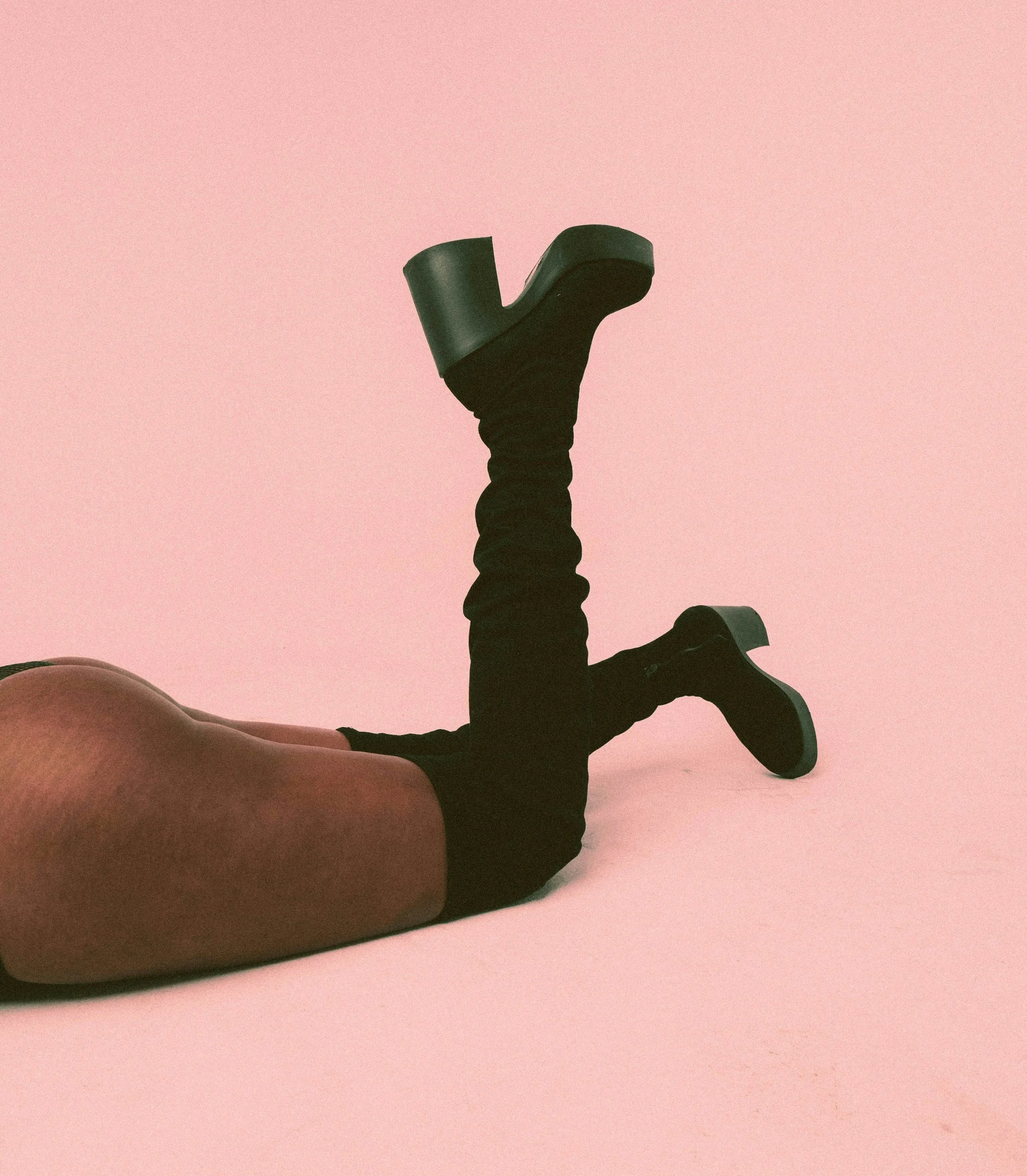 Person lying on the floor with legs raised, wearing black thigh-high boots with chunky heels, on a pink background.