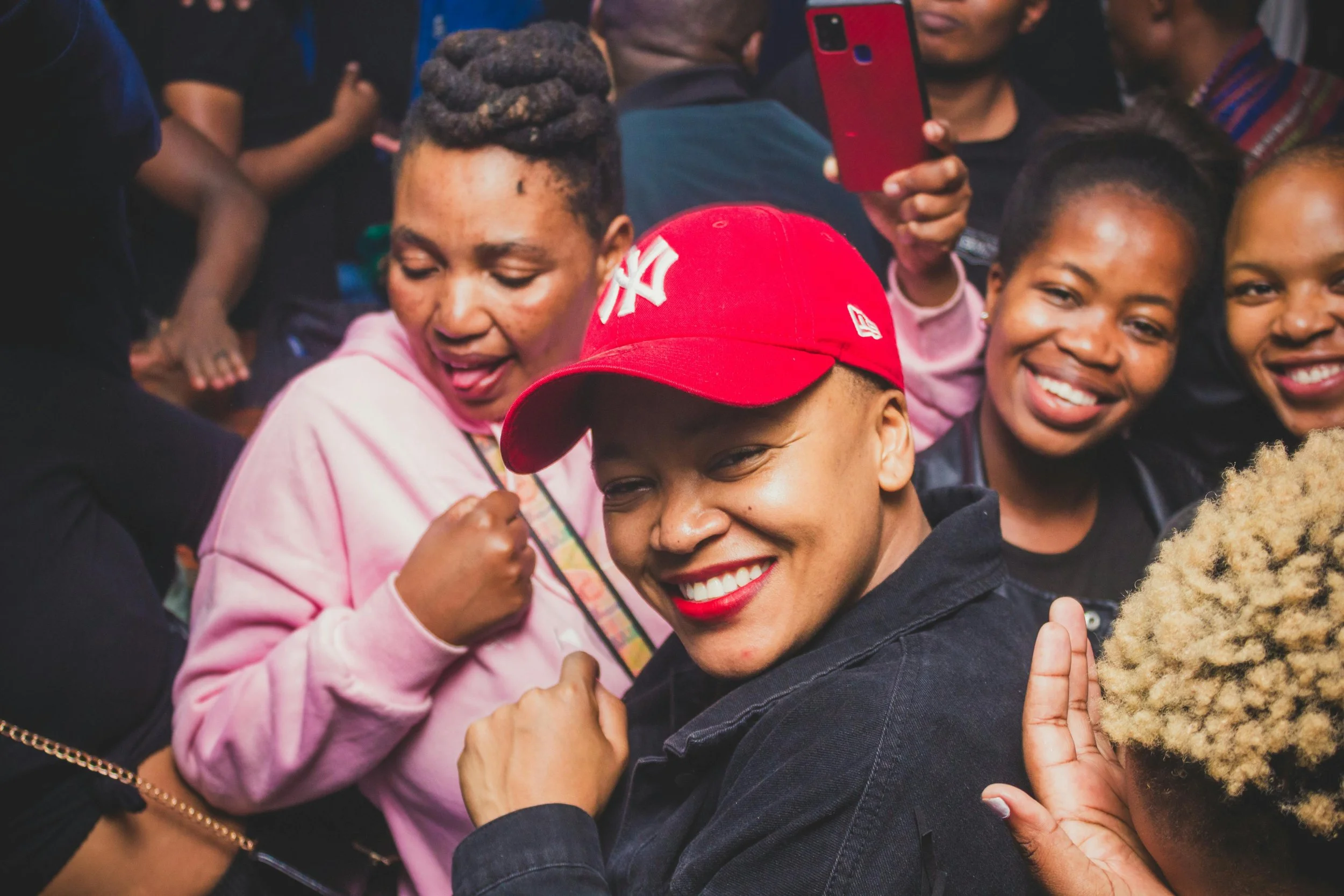 A group of smiling women, with a woman in a red cap at the center, posing for a photo at a crowded event.