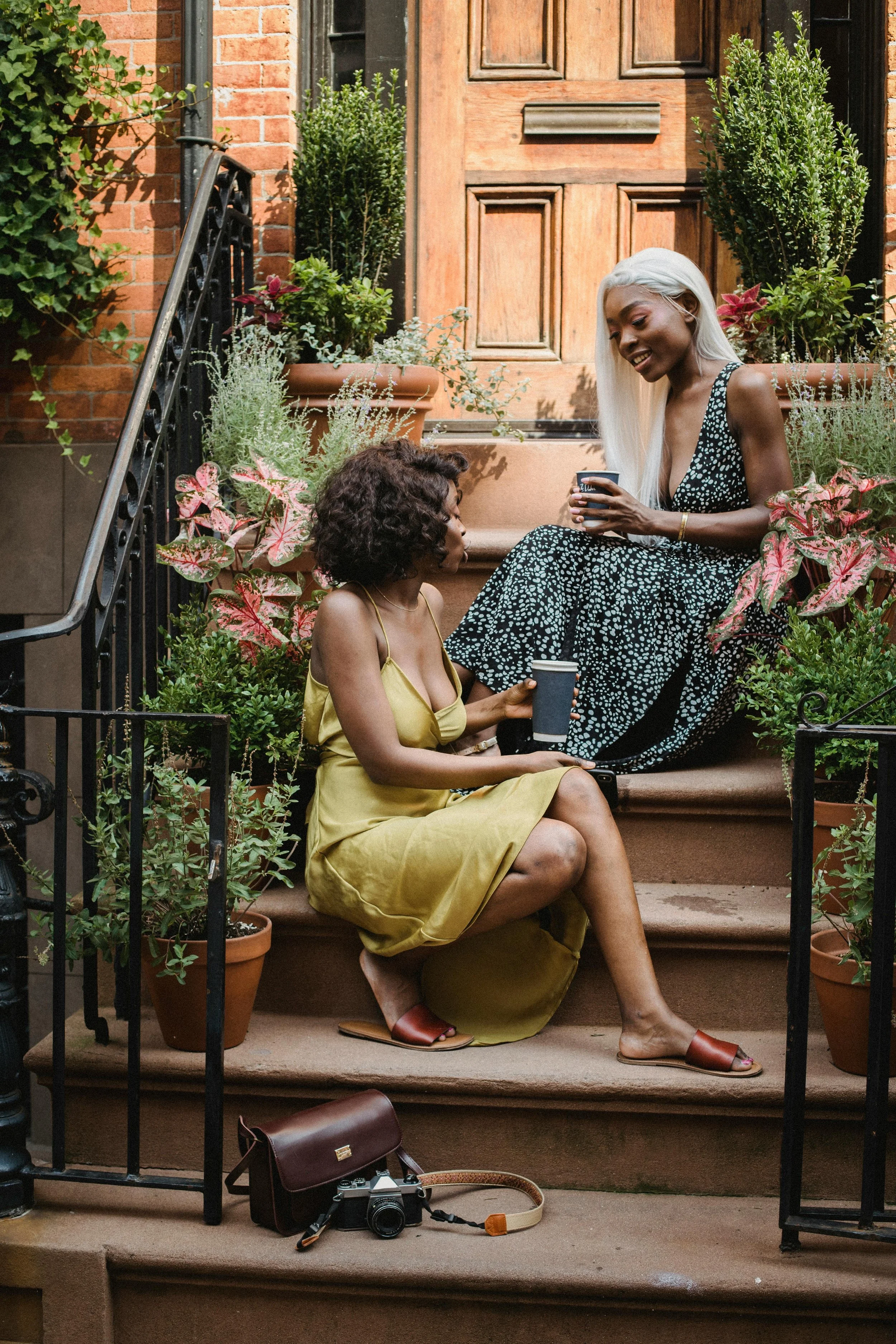 Two women sitting on a stoop outside a brick building, surrounded by potted plants, talking and holding coffee cups, with a camera and bag on the steps.