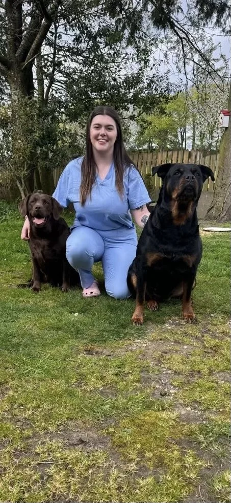 Young woman in blue scrubs kneeling on grass outdoors, flanked by two large dogs, a chocolate Labrador on her left and a Rottweiler on her right, with trees and a wooden fence in the background.