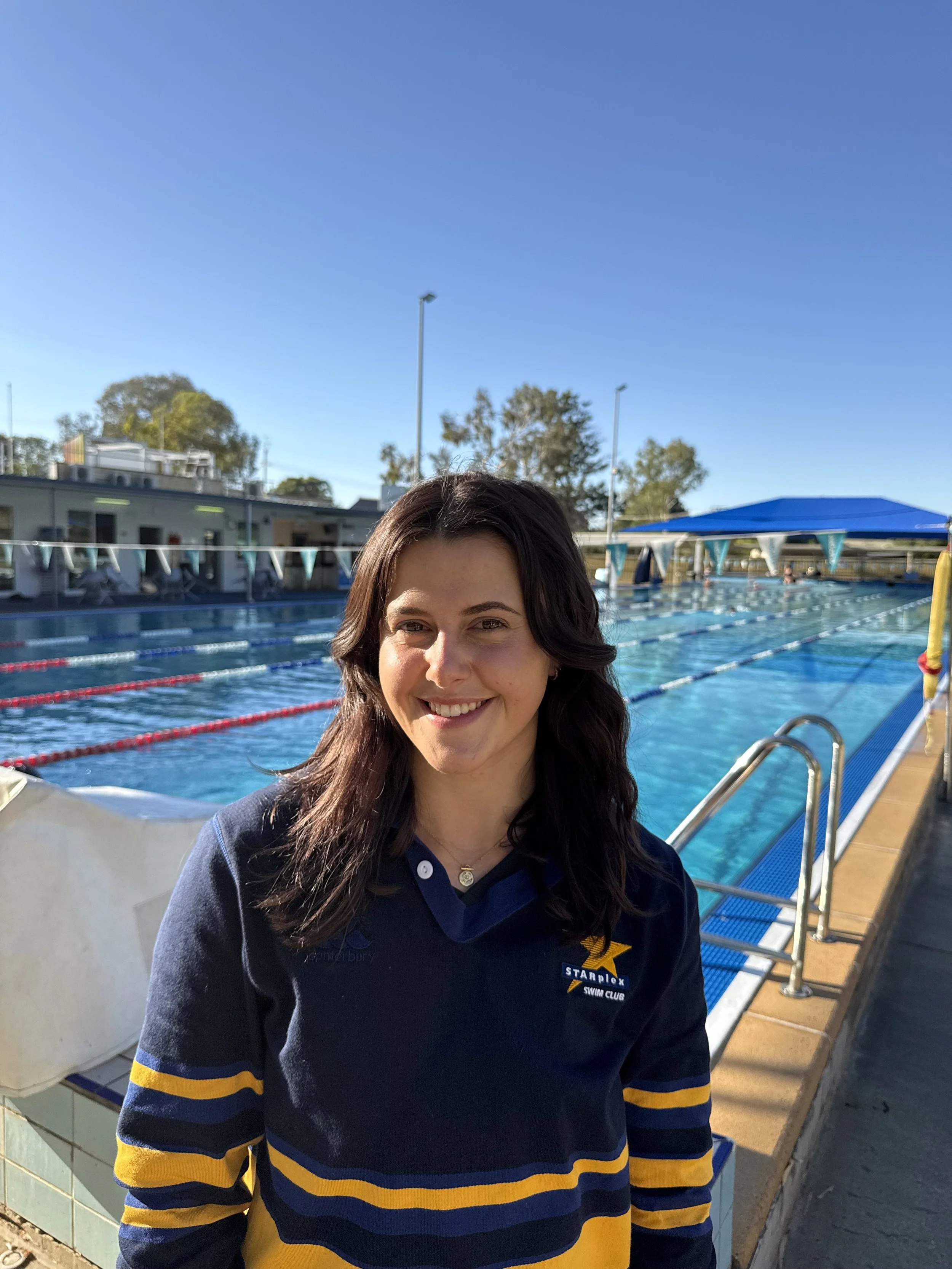 Smiling coach standing near outdoor swimming pool on sunny day, wearing navy and yellow swim club uniform.