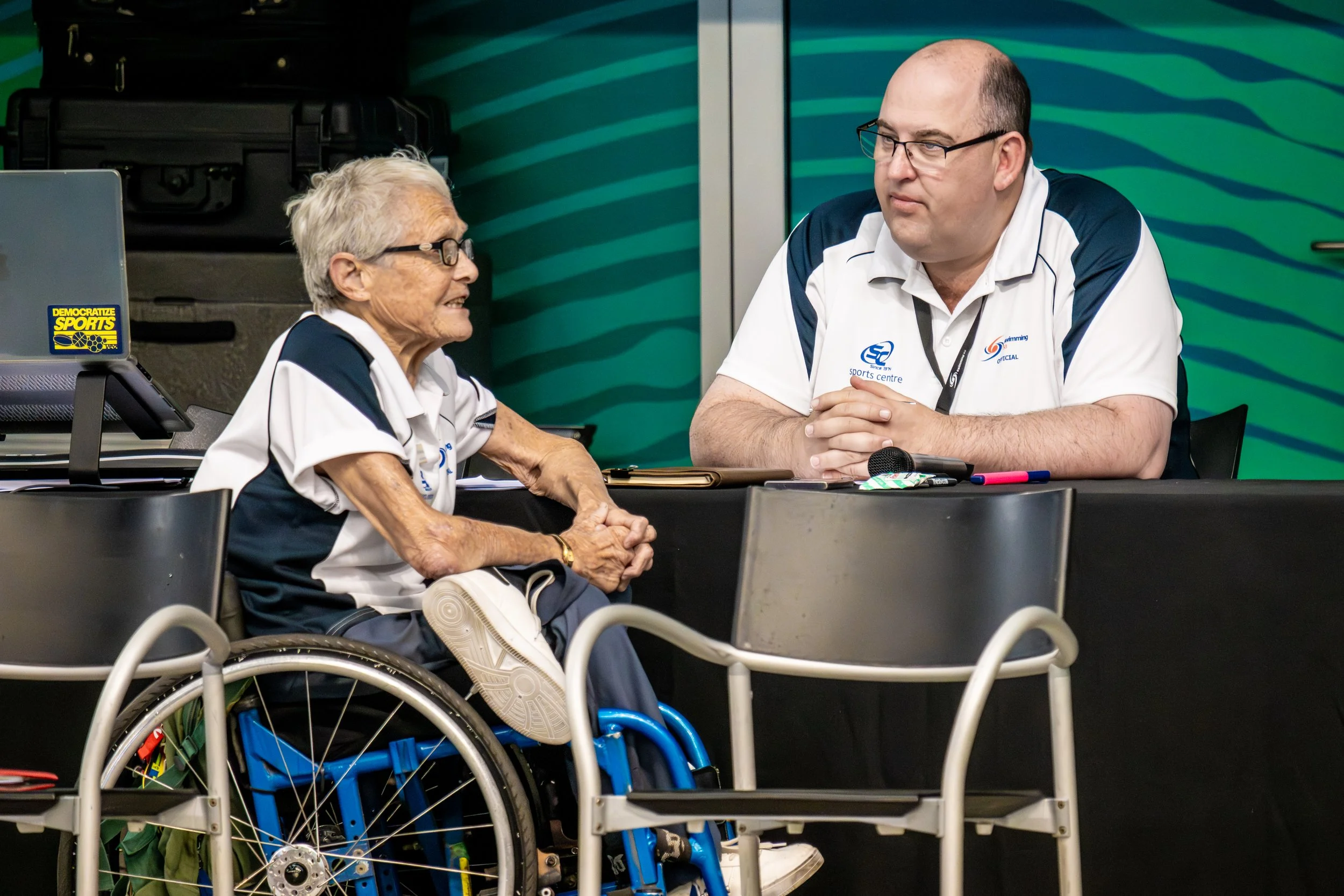 An elderly woman in a wheelchair talking to a man at a table in a sports center.