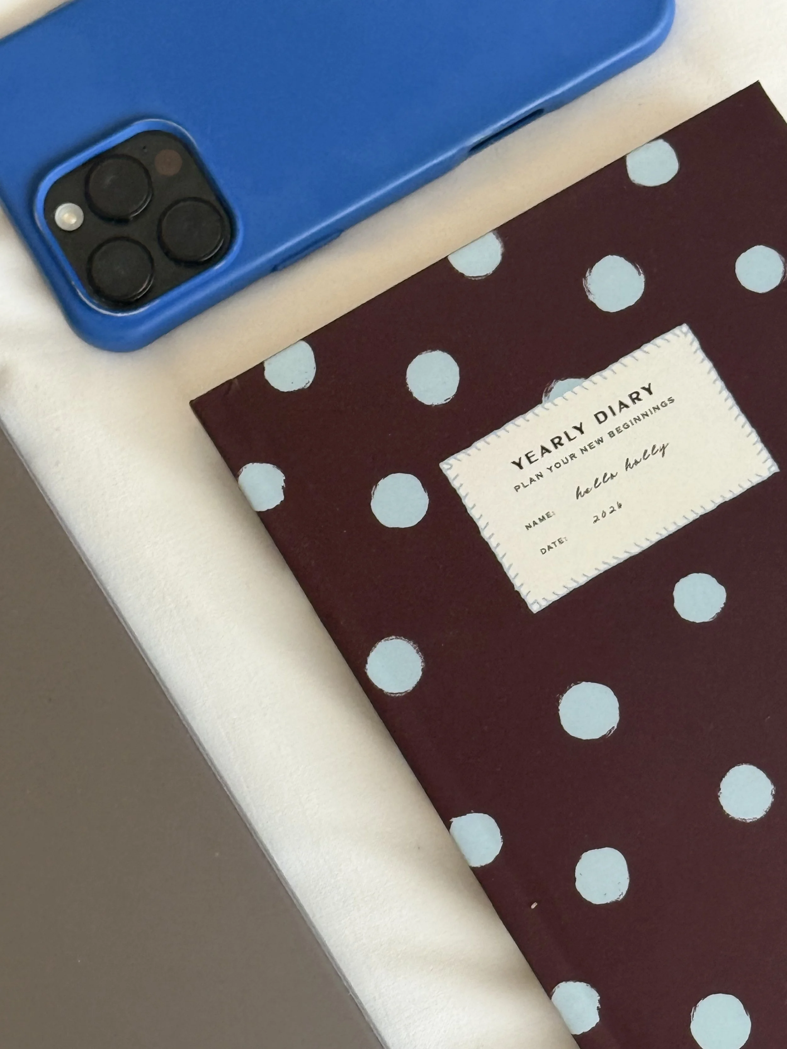 A purple polka dot diary labeled 'Hello Holly Yearly Diary' next to a blue smartphone on a white surface.
