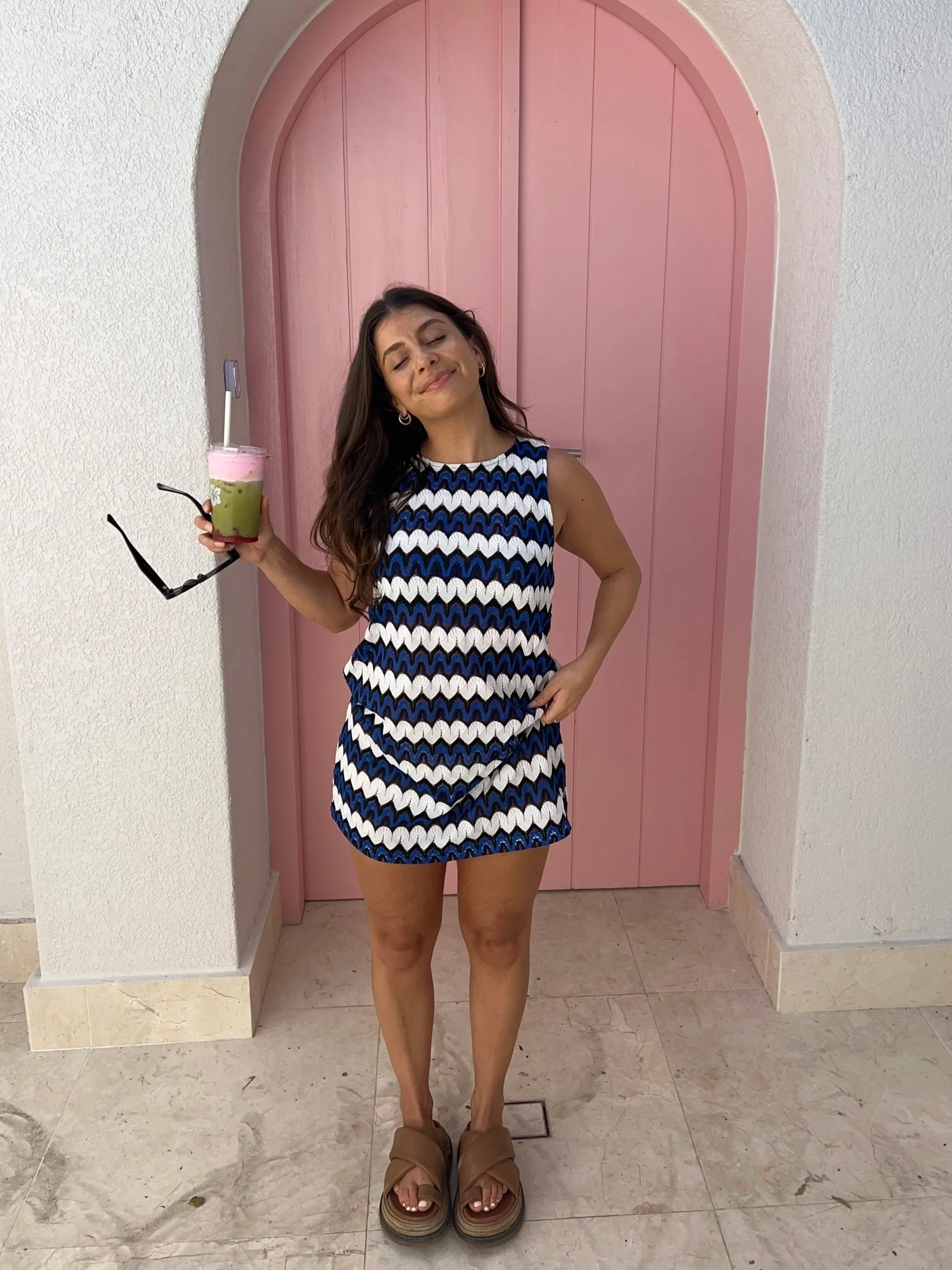 Woman in a blue, black, and white patterned dress standing in front of a pink arched door, holding a green and pink beverage with a straw, smiling with eyes closed, wearing tan sandals.