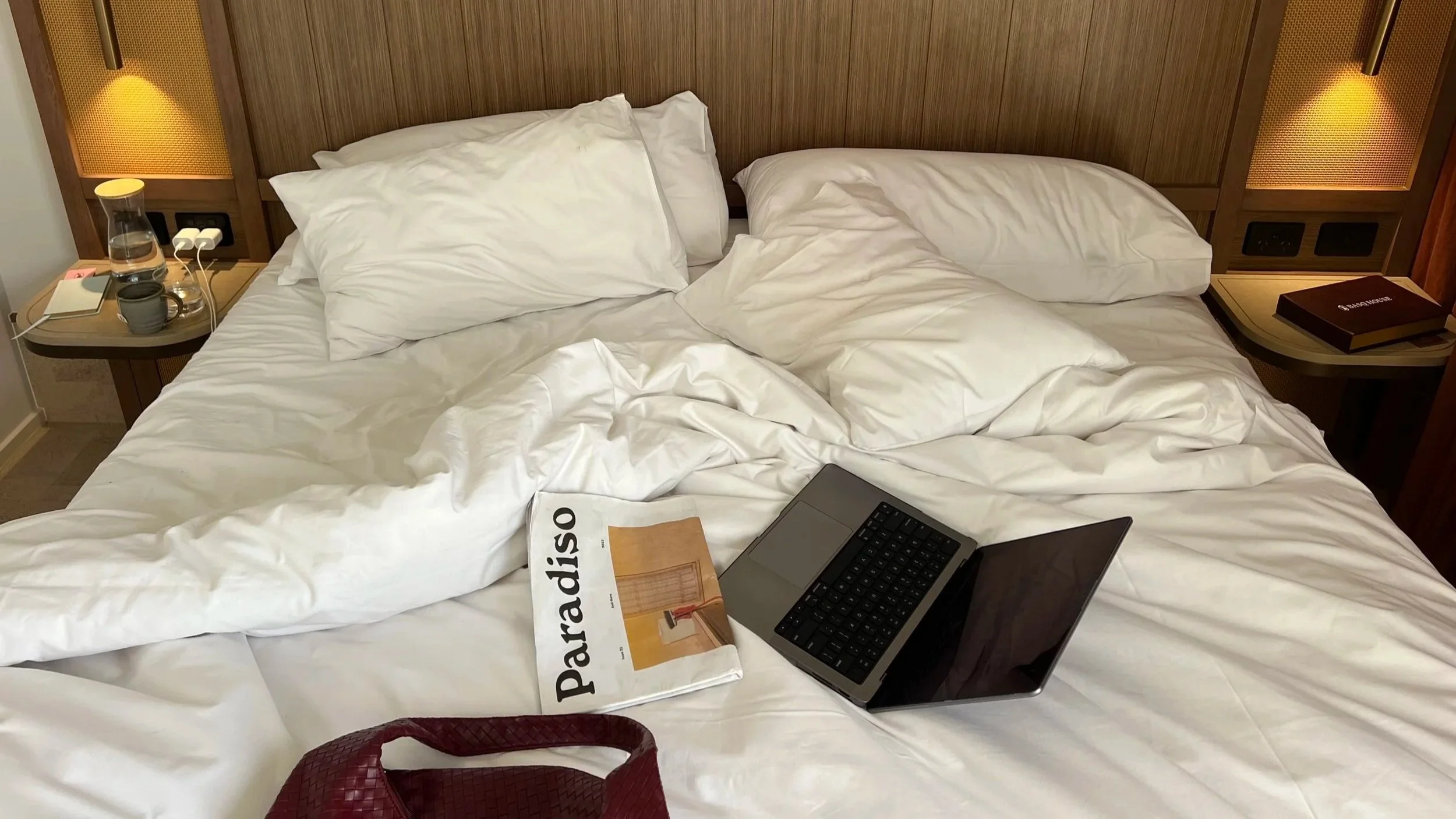 Unmade bed with white linens, two pillows, a laptop, a travel guidebook titled 'Paradiso', a red woven bag, and items on bedside tables including a water bottle, mug, and book.