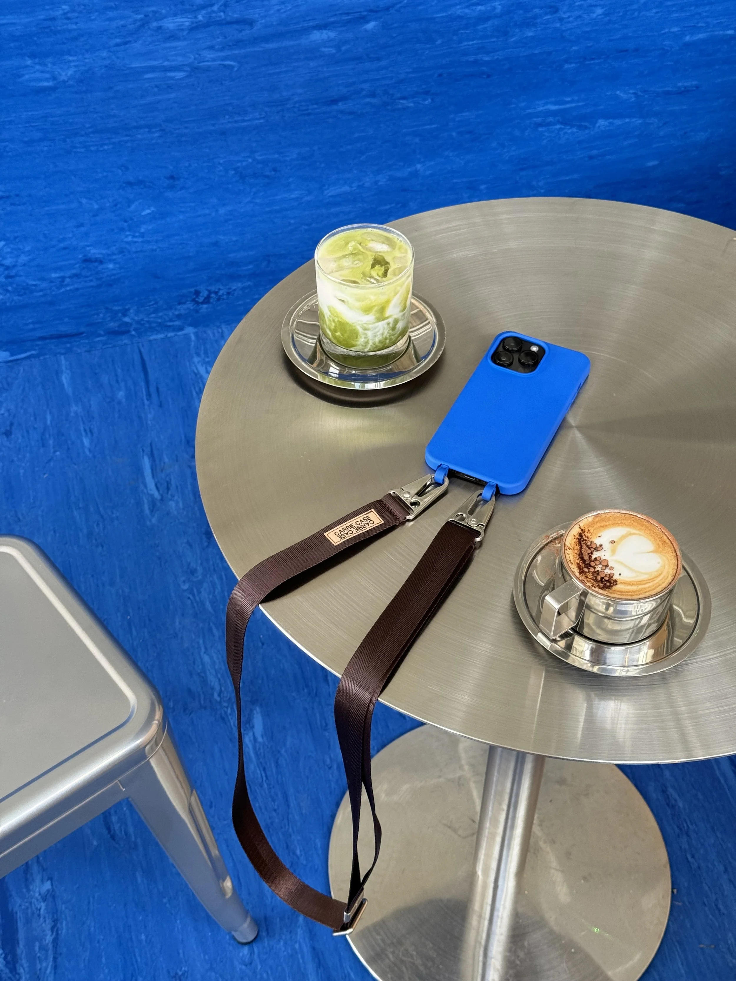 A round metallic table with two cups of coffee, a glass of iced green drink, and a blue smartphone with a brown lanyard attached, set against a bright blue wall.