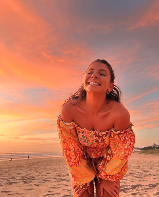 Woman smiling on a beach at sunset, wearing an off-the-shoulder floral dress with long sleeves.