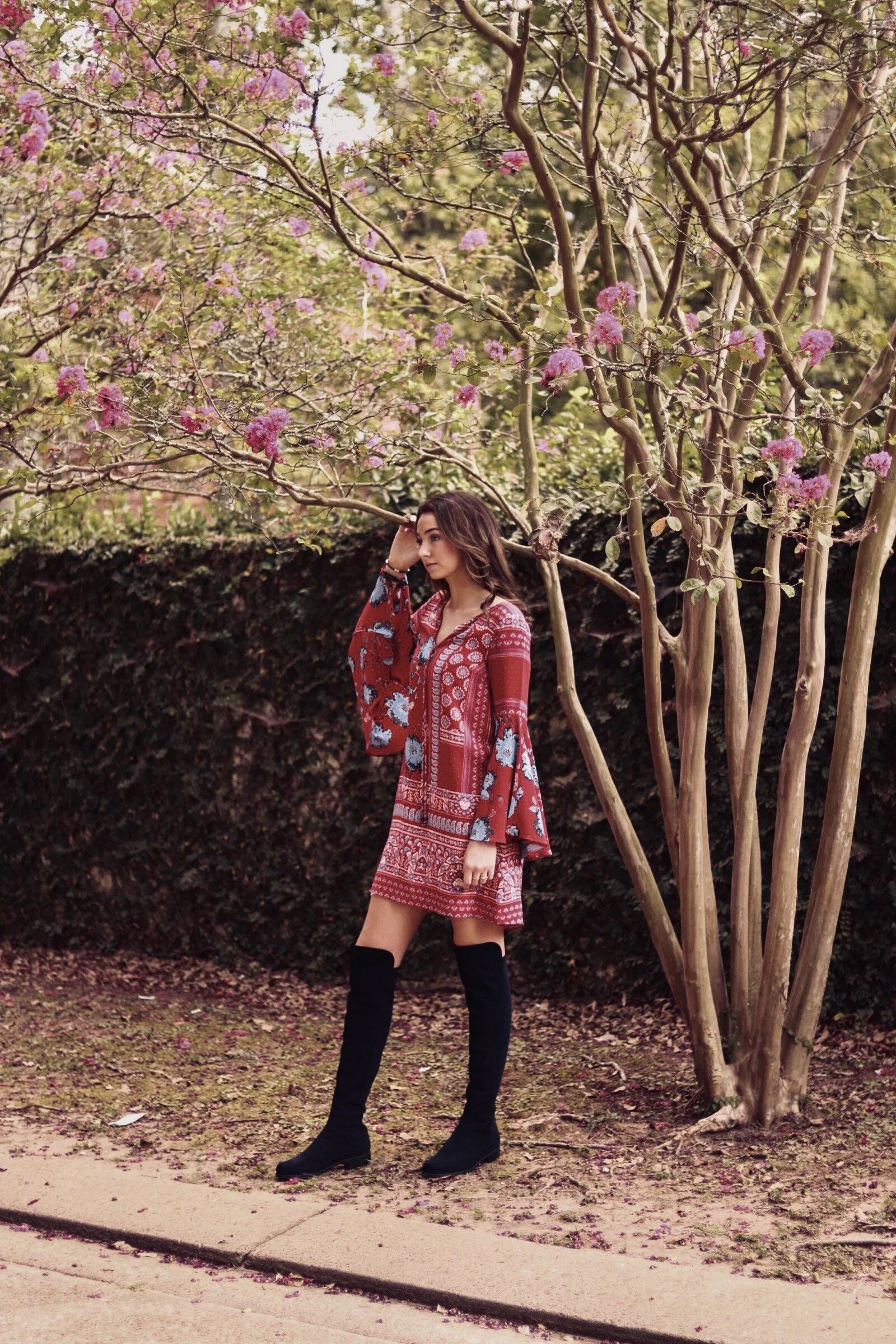 A woman in a red patterned dress and black thigh-high boots posing outdoors near a tree with pink flowers and a hedge in the background.