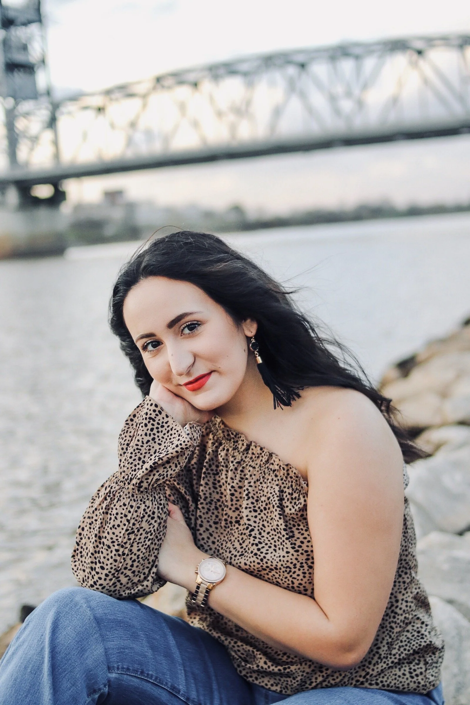 A woman with dark hair and red lipstick sitting on rocks by a river with a bridge in the background.