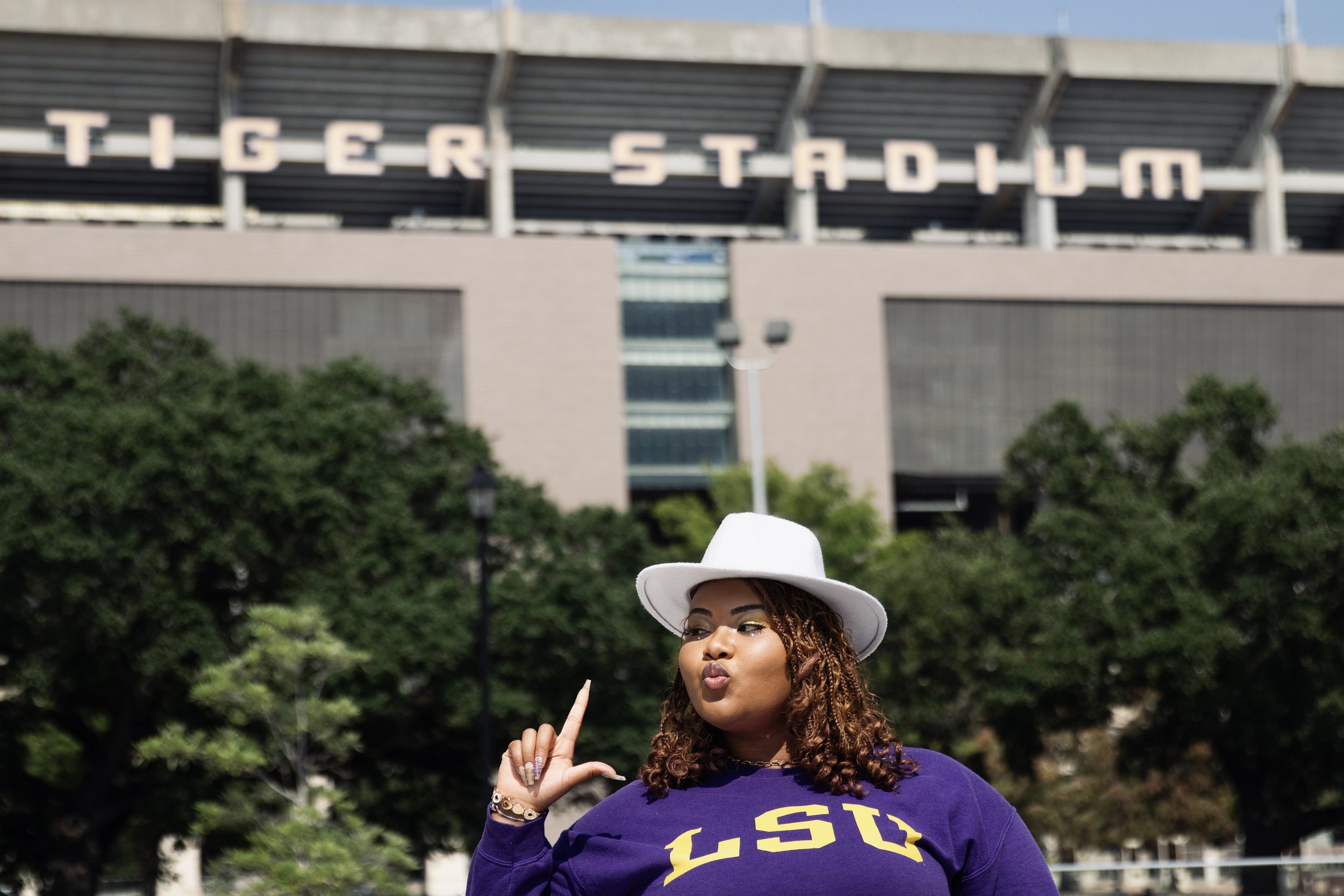 A woman wearing a white hat and purple LSU shirt standing in front of Tiger Stadium, raising one finger in a sign of school spirit.