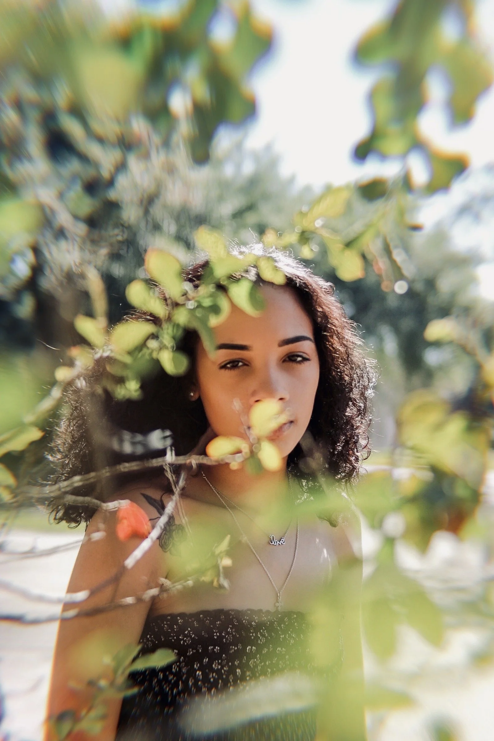 A young woman with curly dark hair and light skin is outdoors among green leaves and branches, with sunlight illuminating her face, wearing a black strapless top with small white polka dots, and necklaces, including one with the word "love."