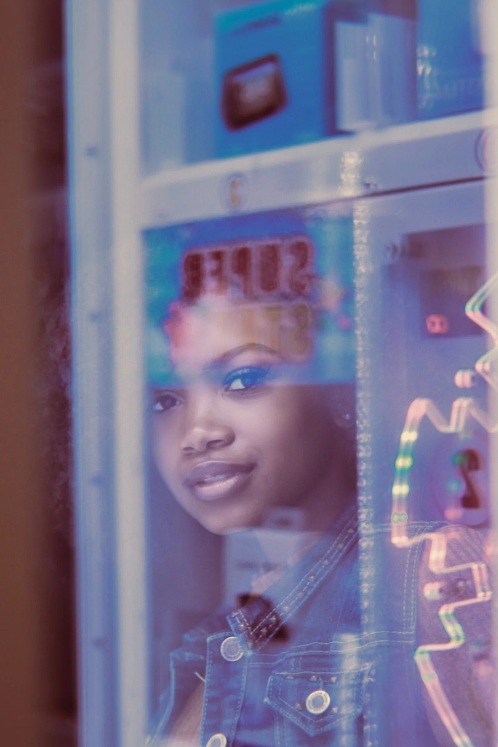 A woman smiling and looking through a glass window at a store refrigerator, with blurred various colorful items and signs behind the glass.