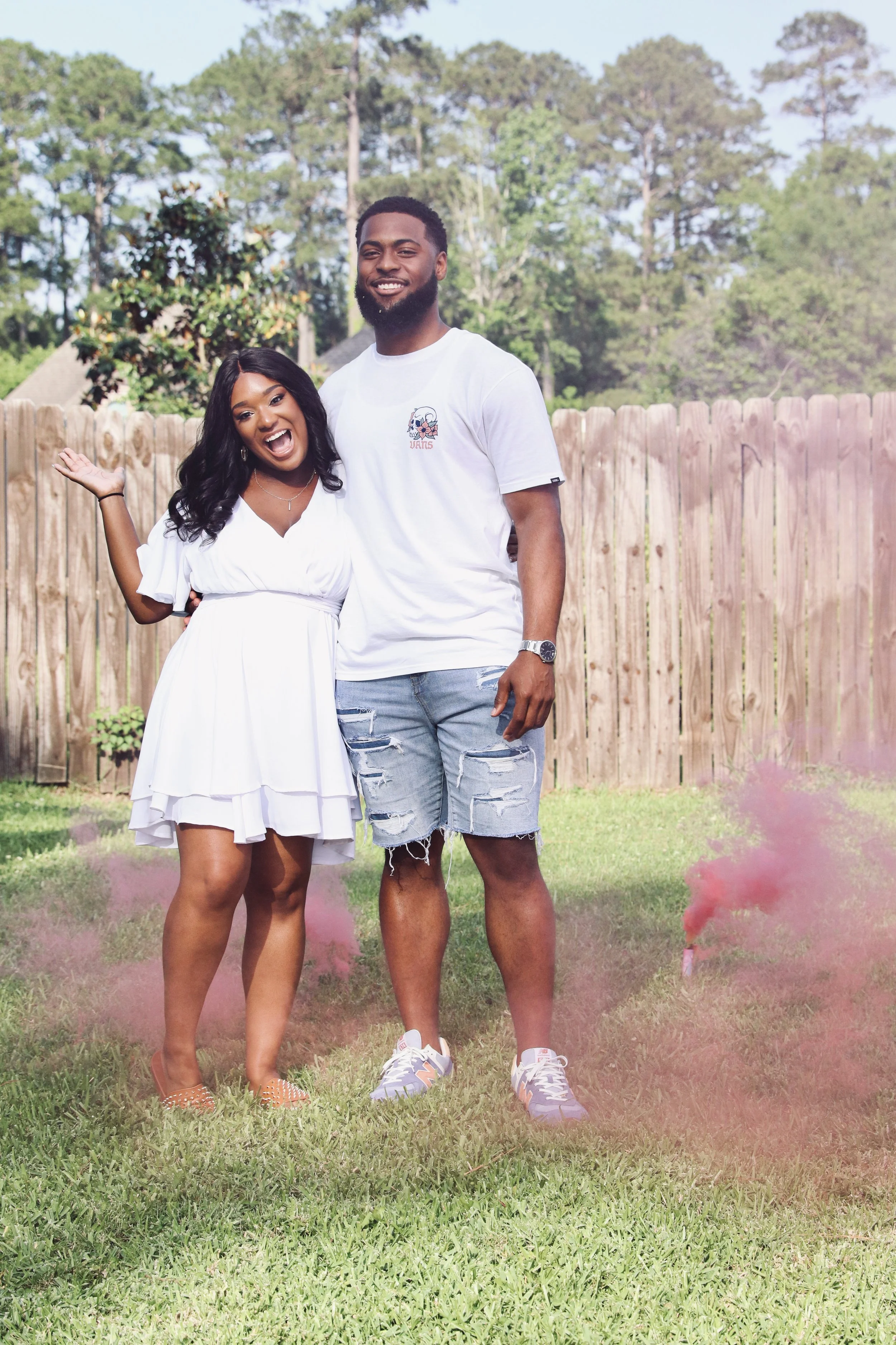 A happy man and woman standing outdoors on a grassy yard, celebrating with pink smoke bombs. The woman is wearing a white dress, and the man is wearing a white t-shirt and ripped denim shorts. Both are smiling, with a wooden fence and trees in the background.