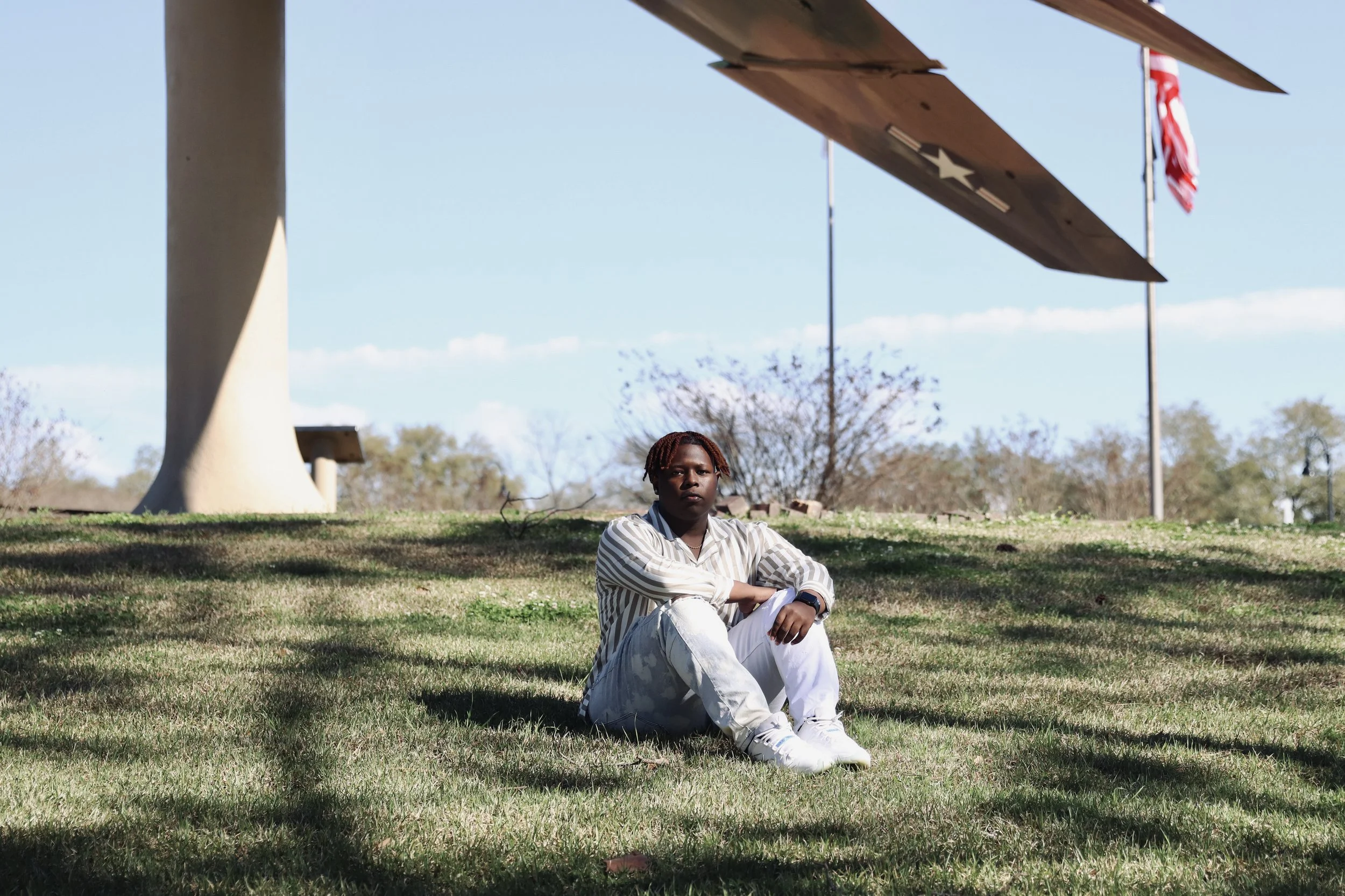A person sitting on the grass in a park-like area under a large landmark with a spacecraft sculpture and American flags in the background.