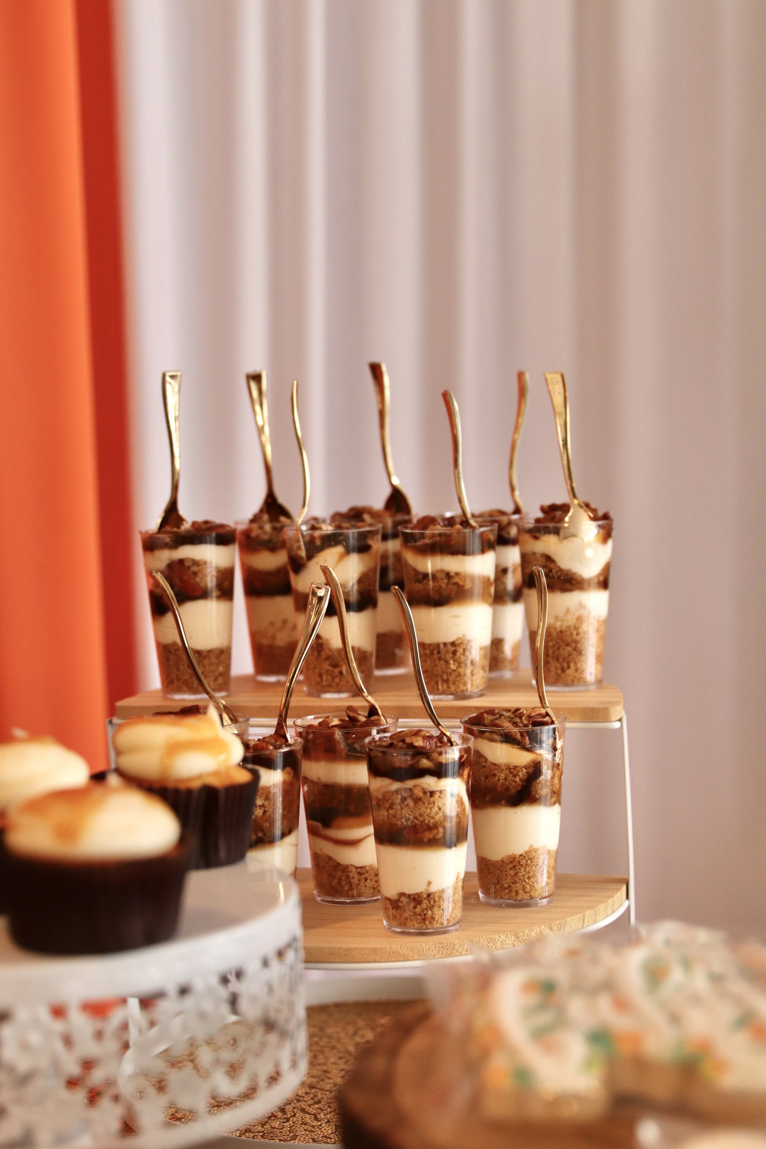 Dessert cups with layered pudding or mousse, topped with chocolate, arranged on wooden and white trays, with blurred desserts in the foreground.