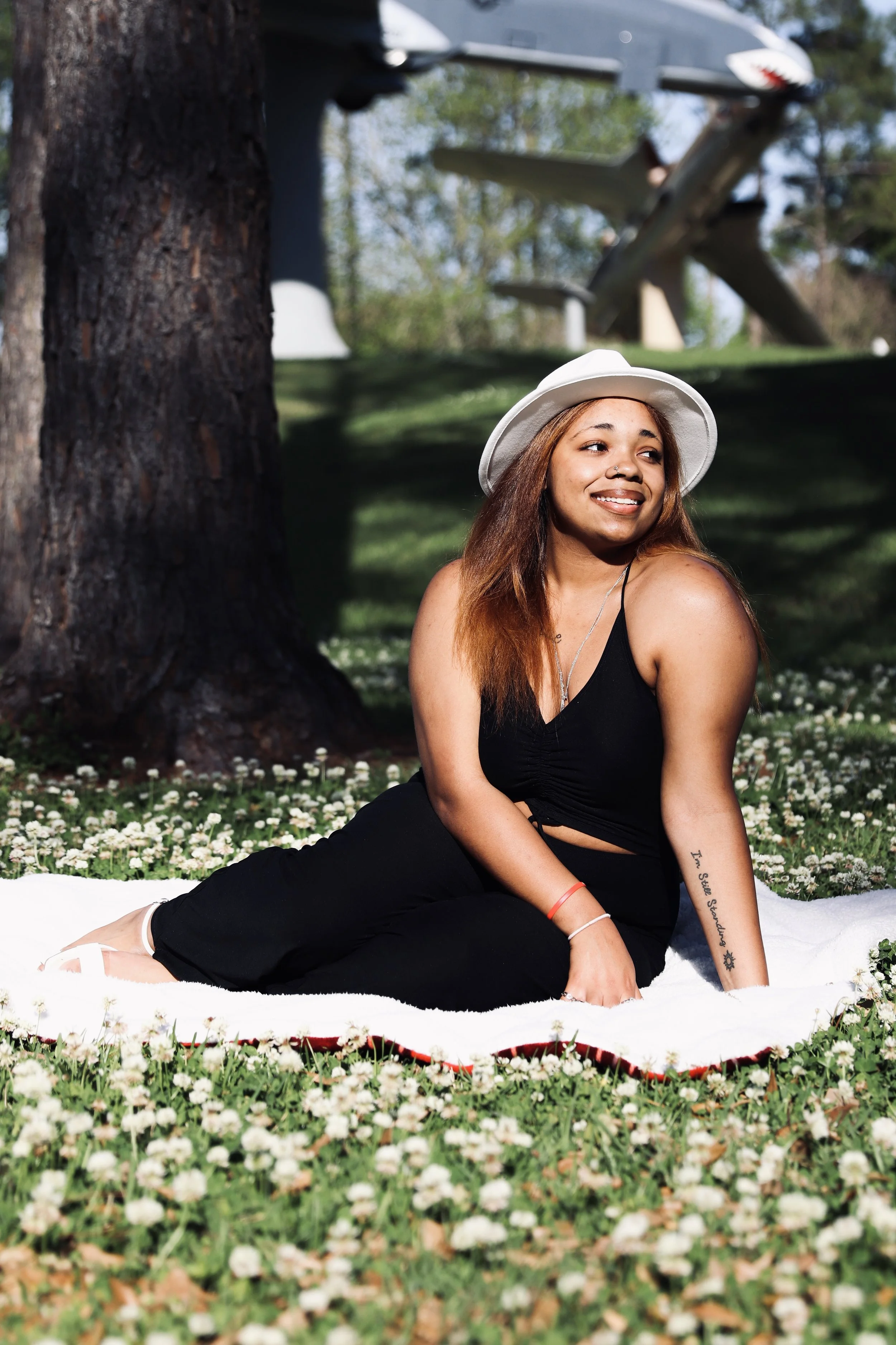 A woman with reddish-brown hair, wearing a white hat and black outfit, sitting on a white blanket on a grassy area with small white flowers, smiling and looking to the side. There is a large tree trunk nearby and airplanes in the background, under a clear sky.
