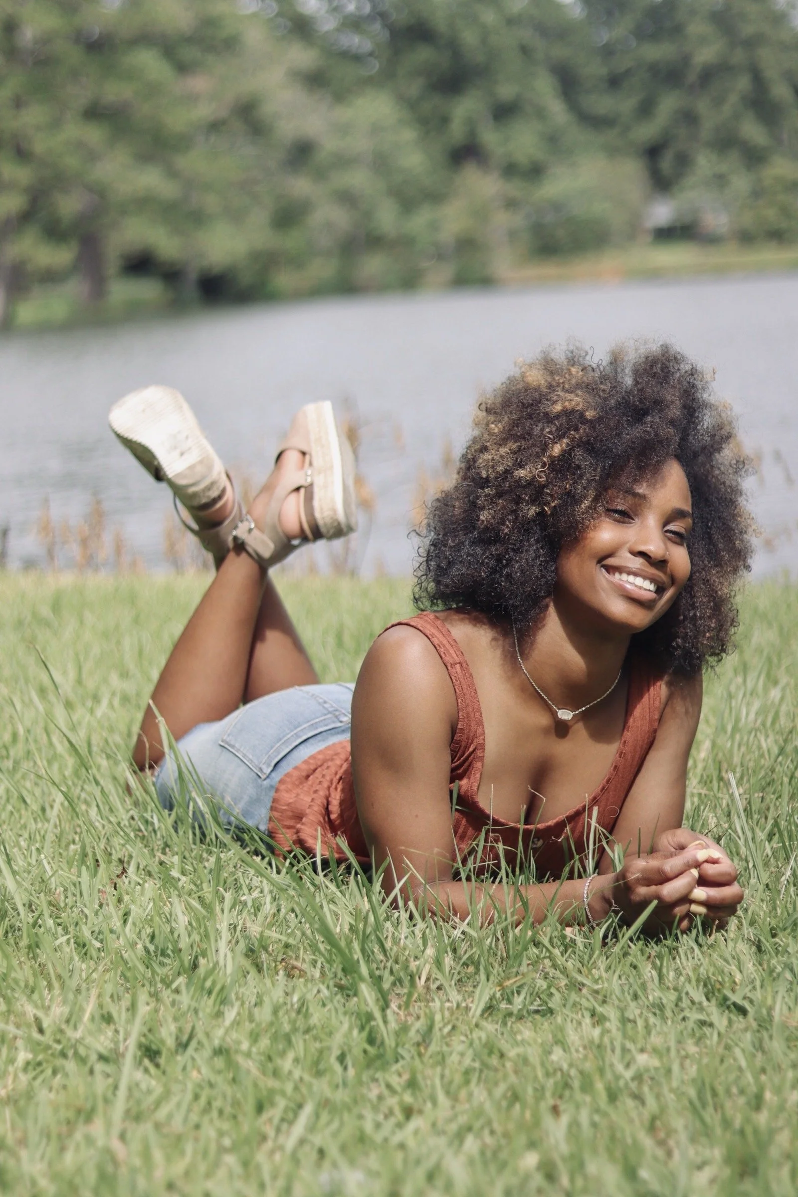 A woman with curly hair lying on the grass near a lake, smiling.