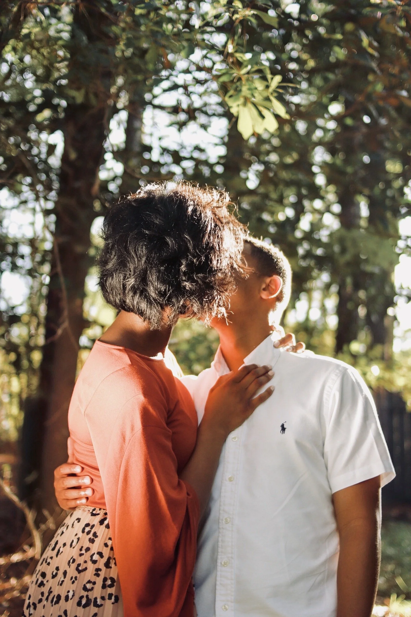 A couple sharing a kiss outdoors, with trees and sunlight in the background.