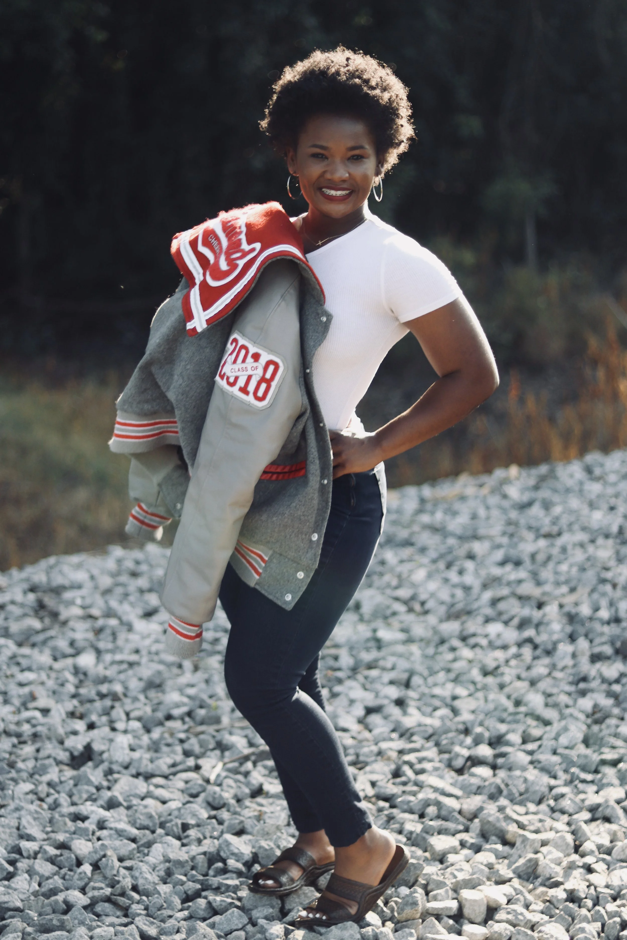 A woman standing outdoors on rocky ground, smiling at the camera, wearing a white t-shirt, black jeans, and brown slides, with a grey and red letterman jacket draped over her shoulder.