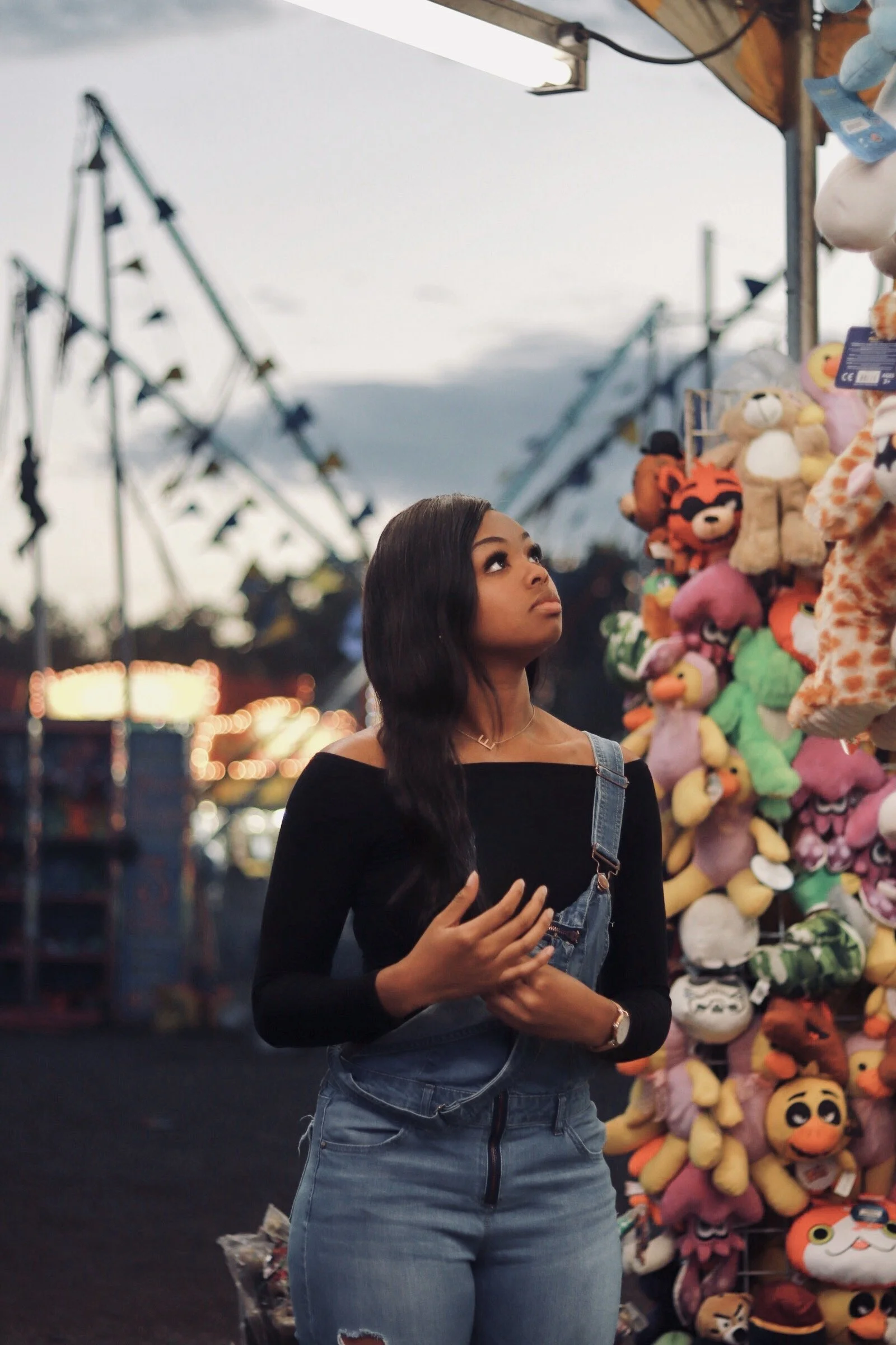 A woman at a carnival or amusement park during dusk, looking up at plush toys hanging on a prize stall, with rides and lights blurred in the background.