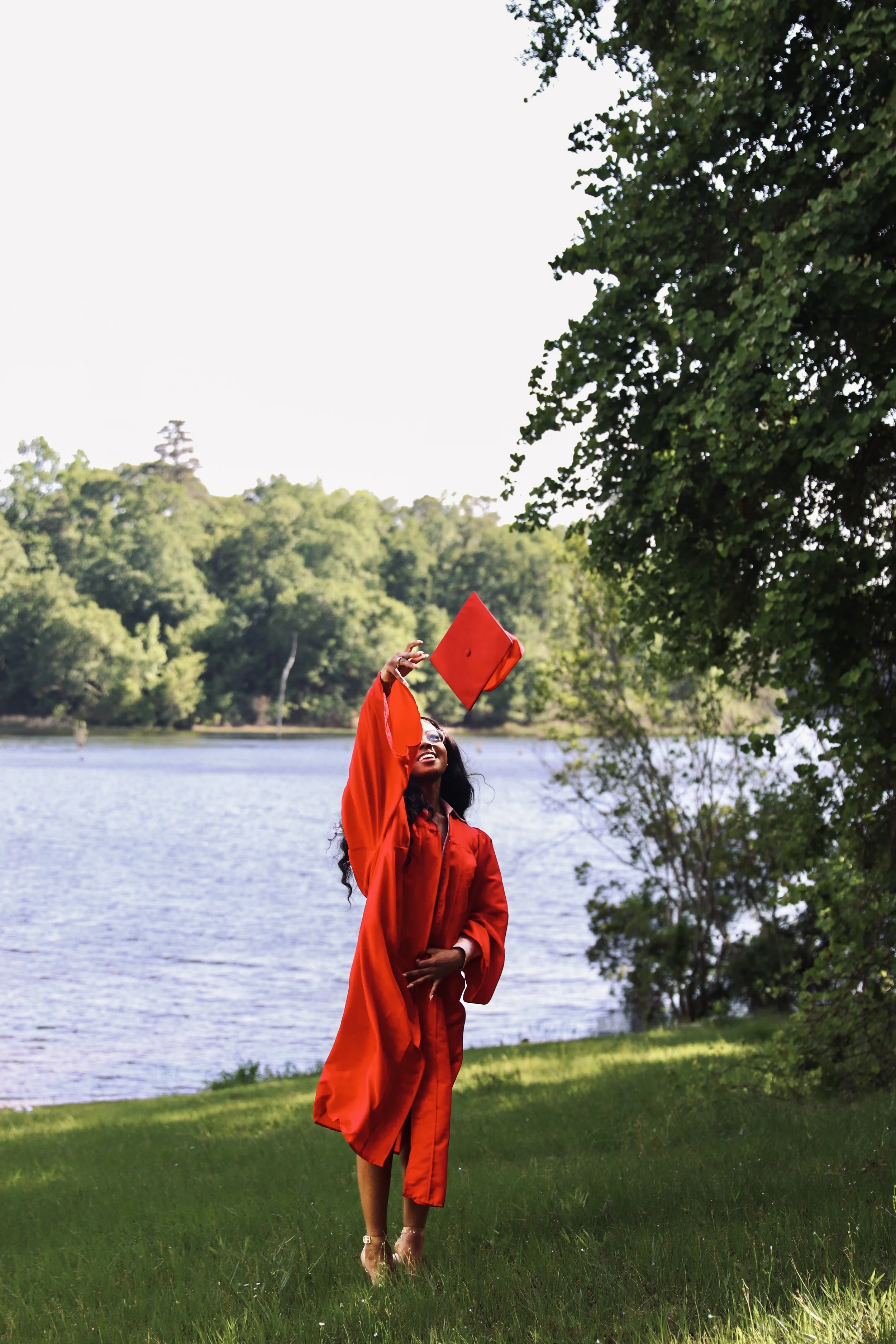 A woman in a red graduation gown and cap taking a selfie outdoors by a lake, with trees in the background.