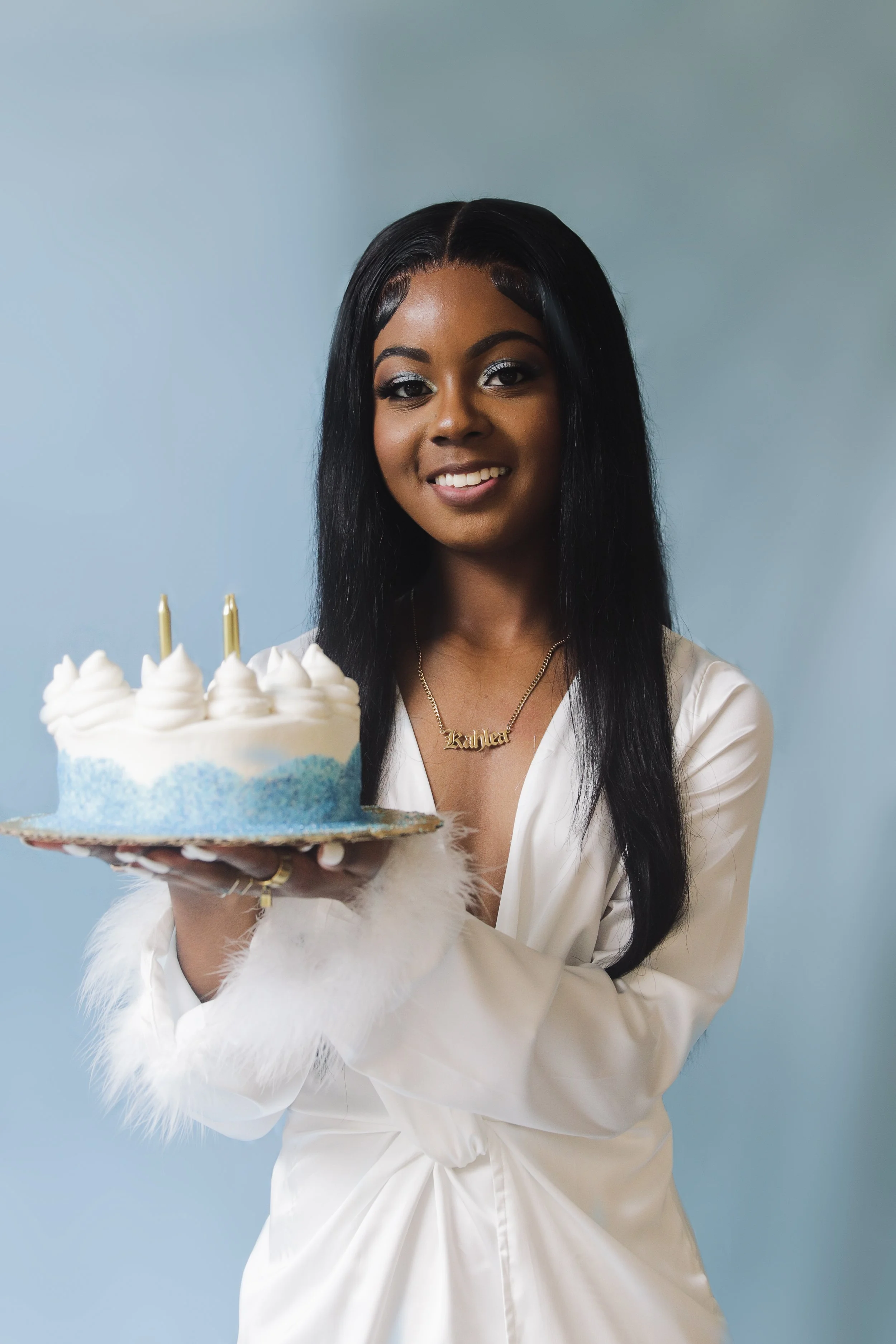 A woman with long black hair and gold jewelry holding a birthday cake with two candles, smiling against a light blue background.