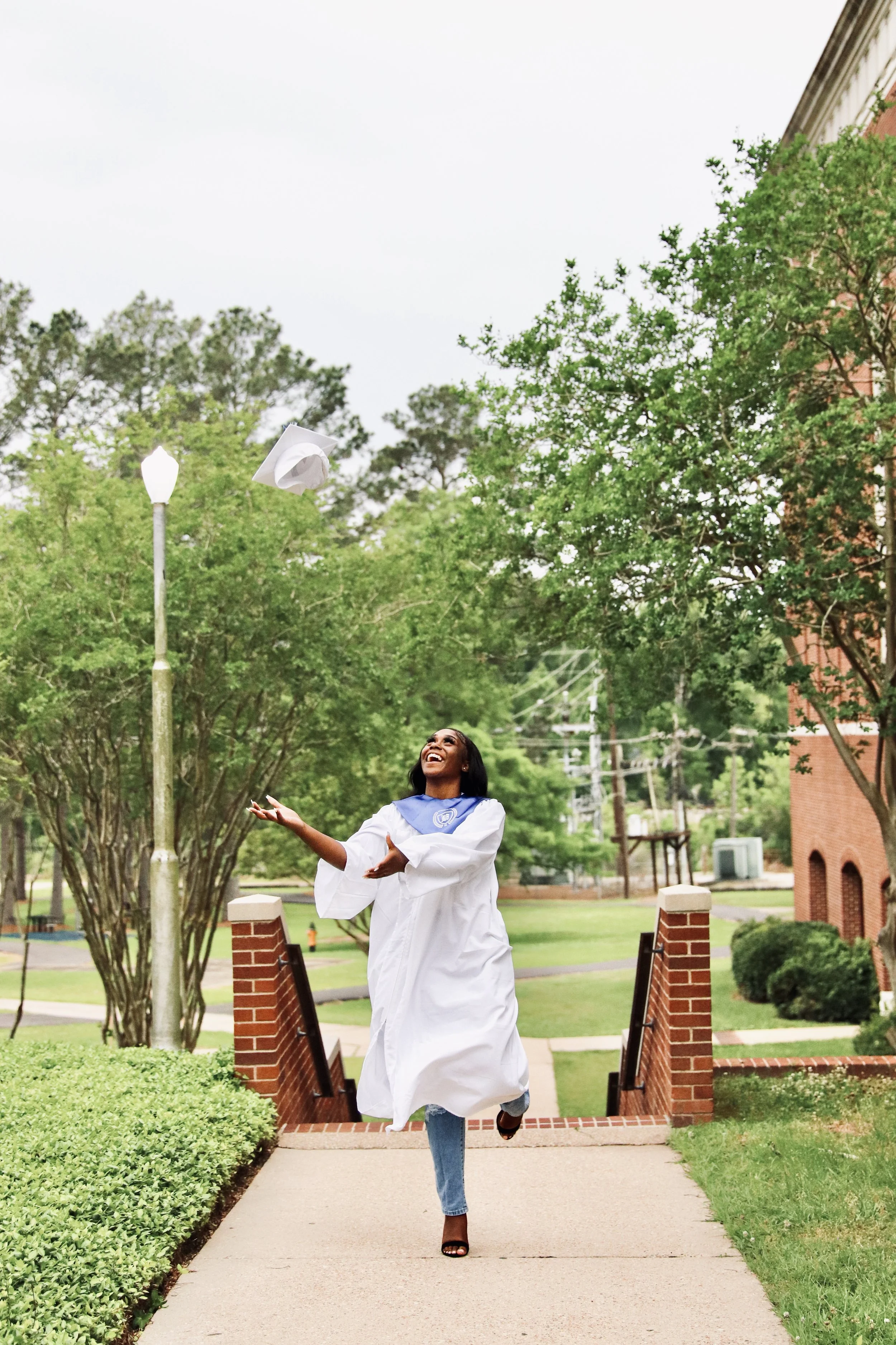 A young woman in a white graduation gown and blue stole celebrating outdoors on a college campus, tossing her cap in the air with a joyful expression.