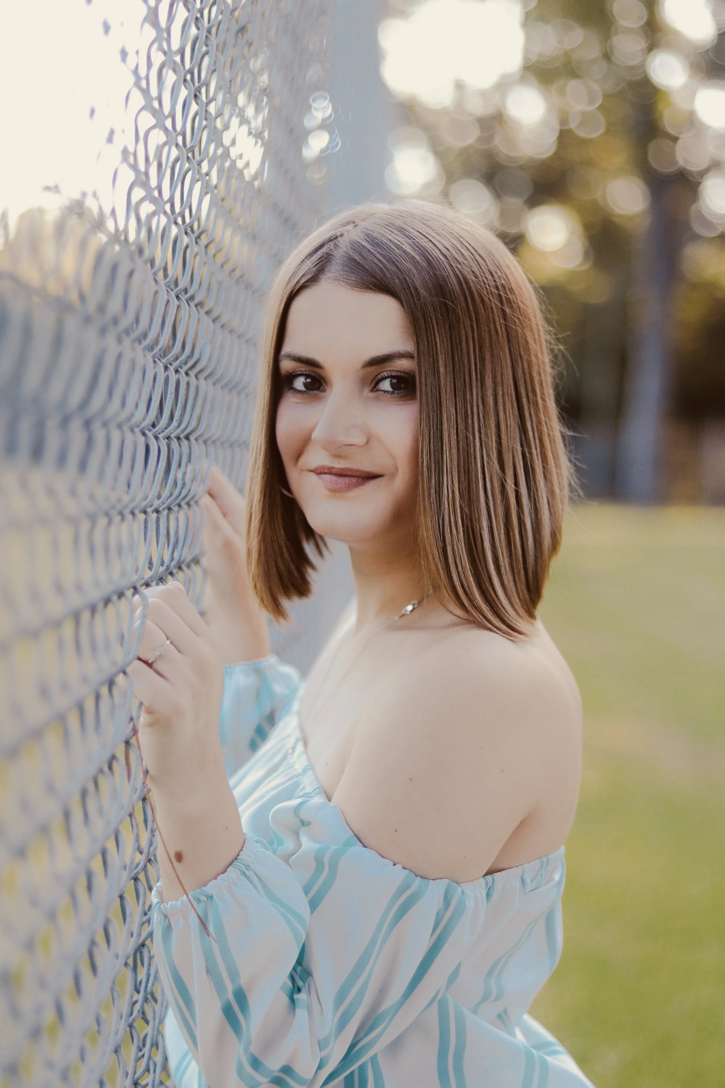 A young woman with shoulder-length brown hair, smiling softly, stands next to a metal chain-link fence outdoors during sunset, wearing an off-shoulder blue and white striped top.