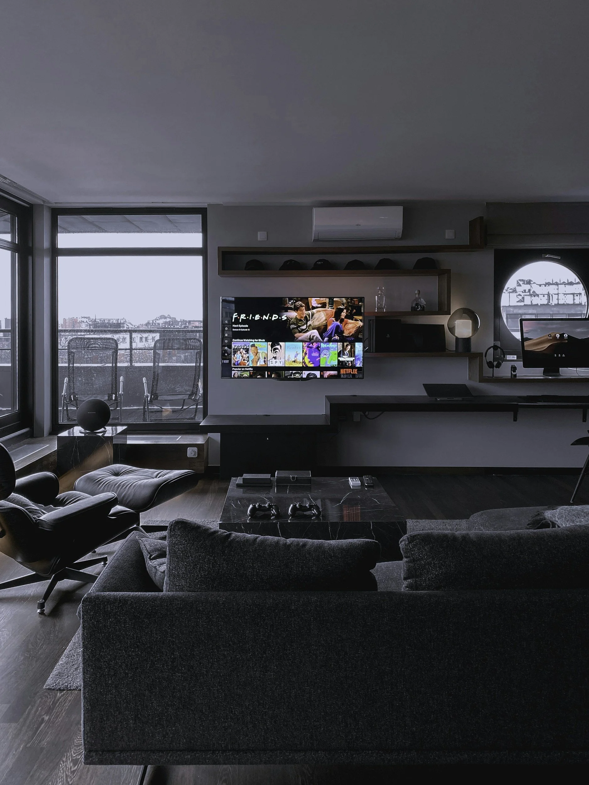 Modern living room with a large window, television, and ergonomic chair, overlooking an outdoor balcony and cityscape.