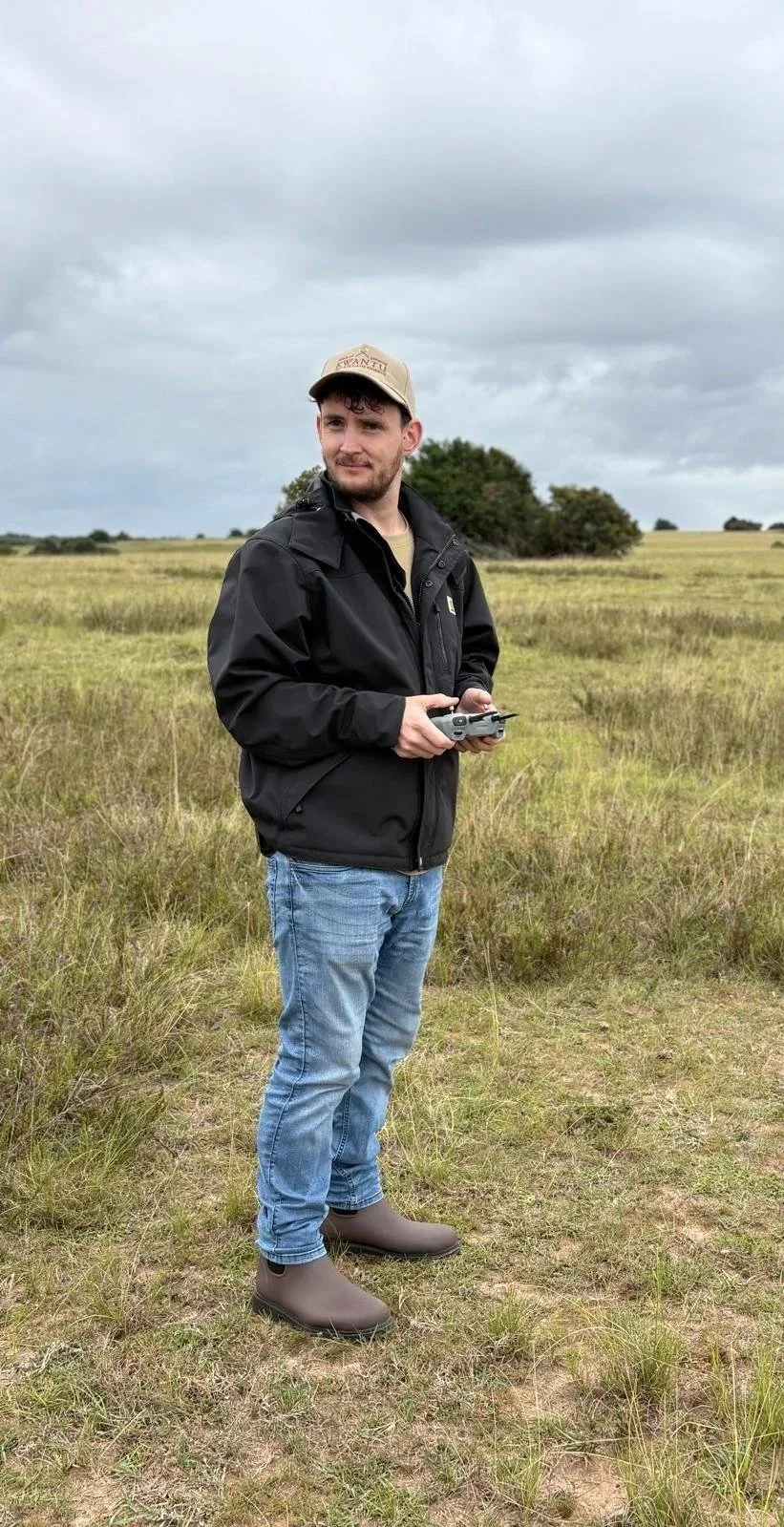 A man standing in an open field holding a drone remote control, wearing a black jacket, jeans, waterproof boots, and a beige cap, with cloudy skies overhead.