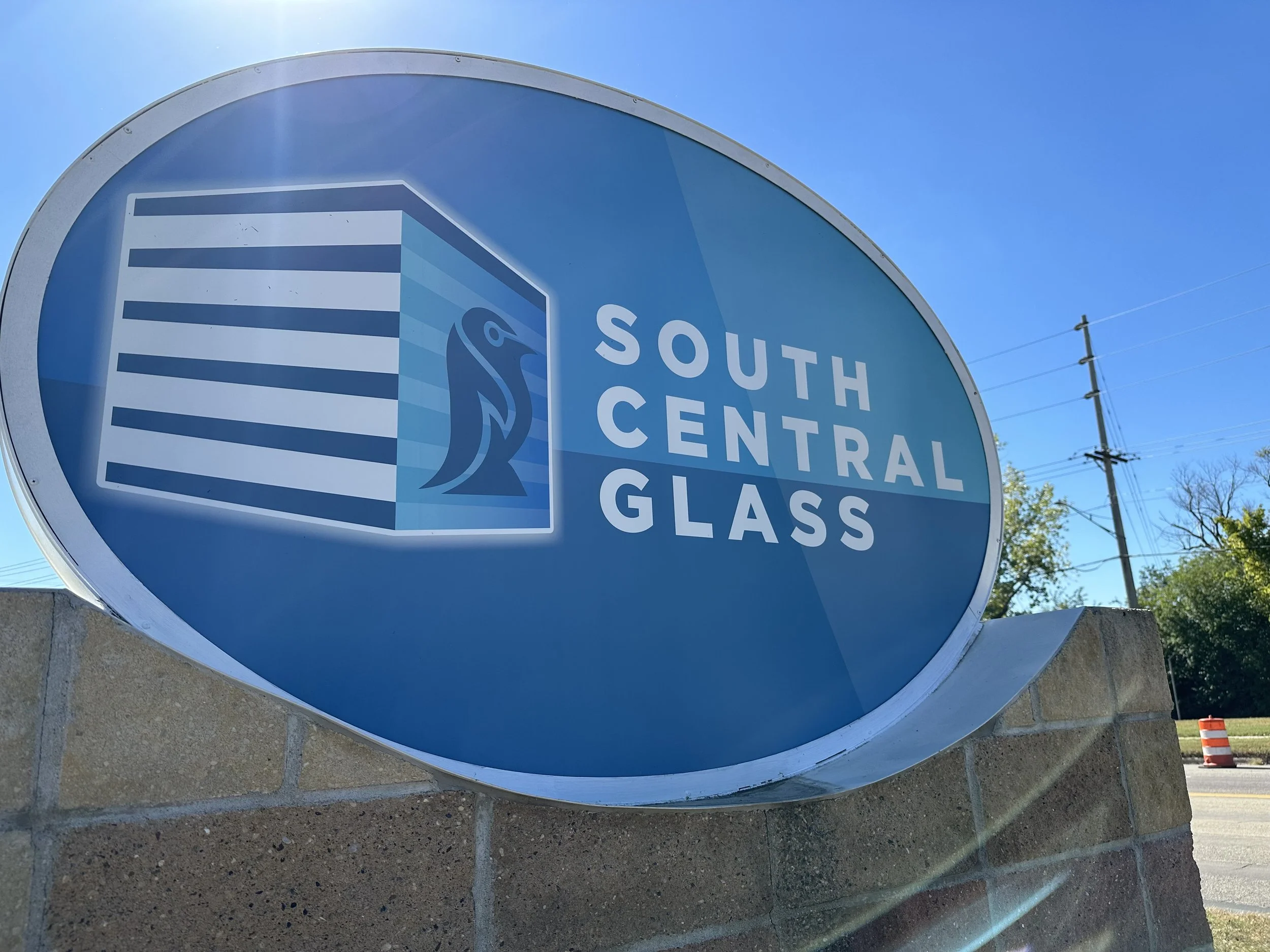 Sign for South Central Glass with a logo featuring a stylized bird and a building with horizontal lines, on a brick base against a clear blue sky.