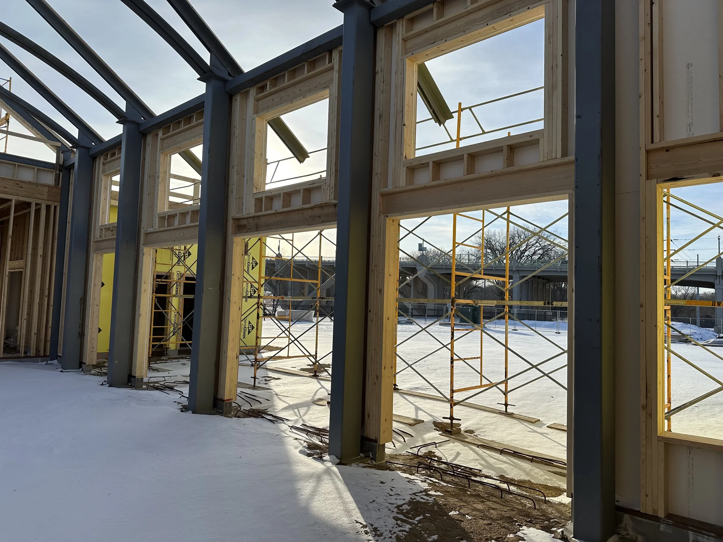 Construction site of a building with wooden framing and large window openings, scaffolding outside, snow on the ground, and a bridge in the background.