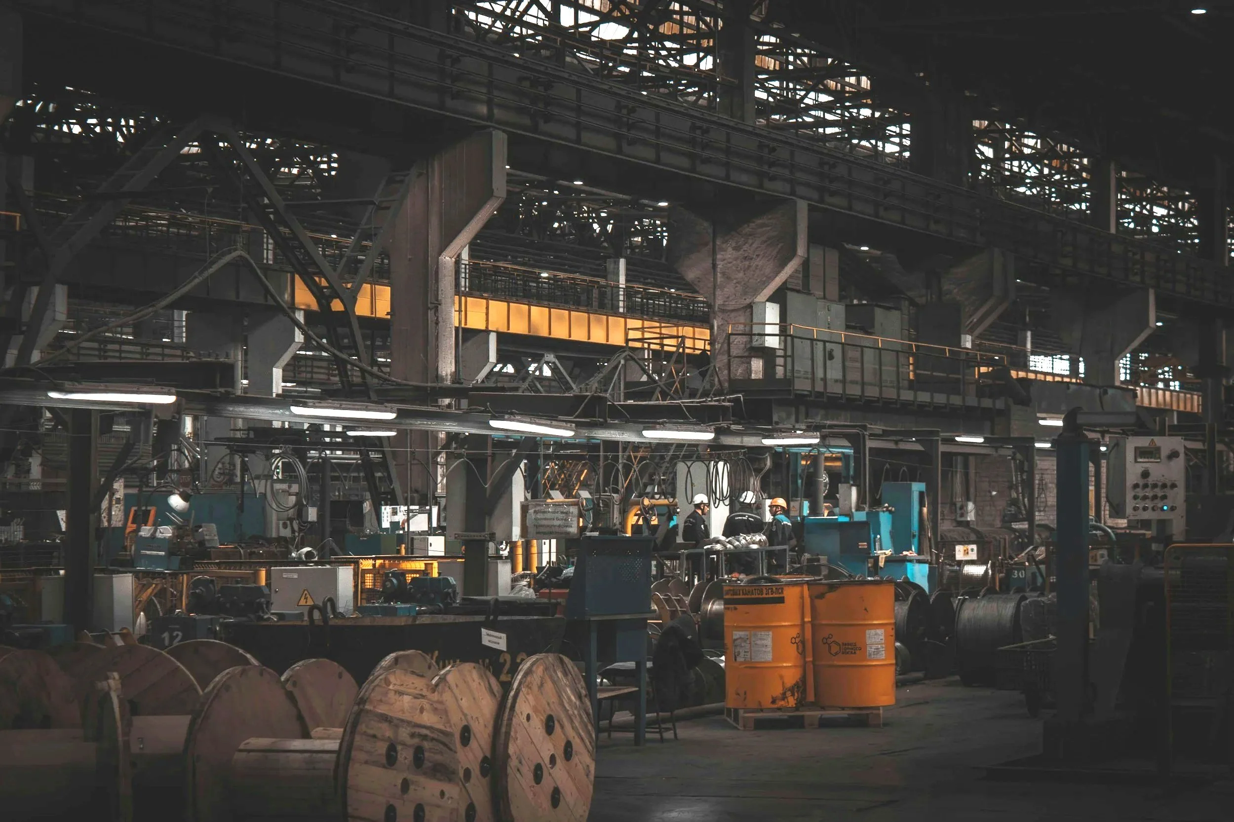 Interior of a large industrial factory with machinery, cable reels, and workers wearing helmets.