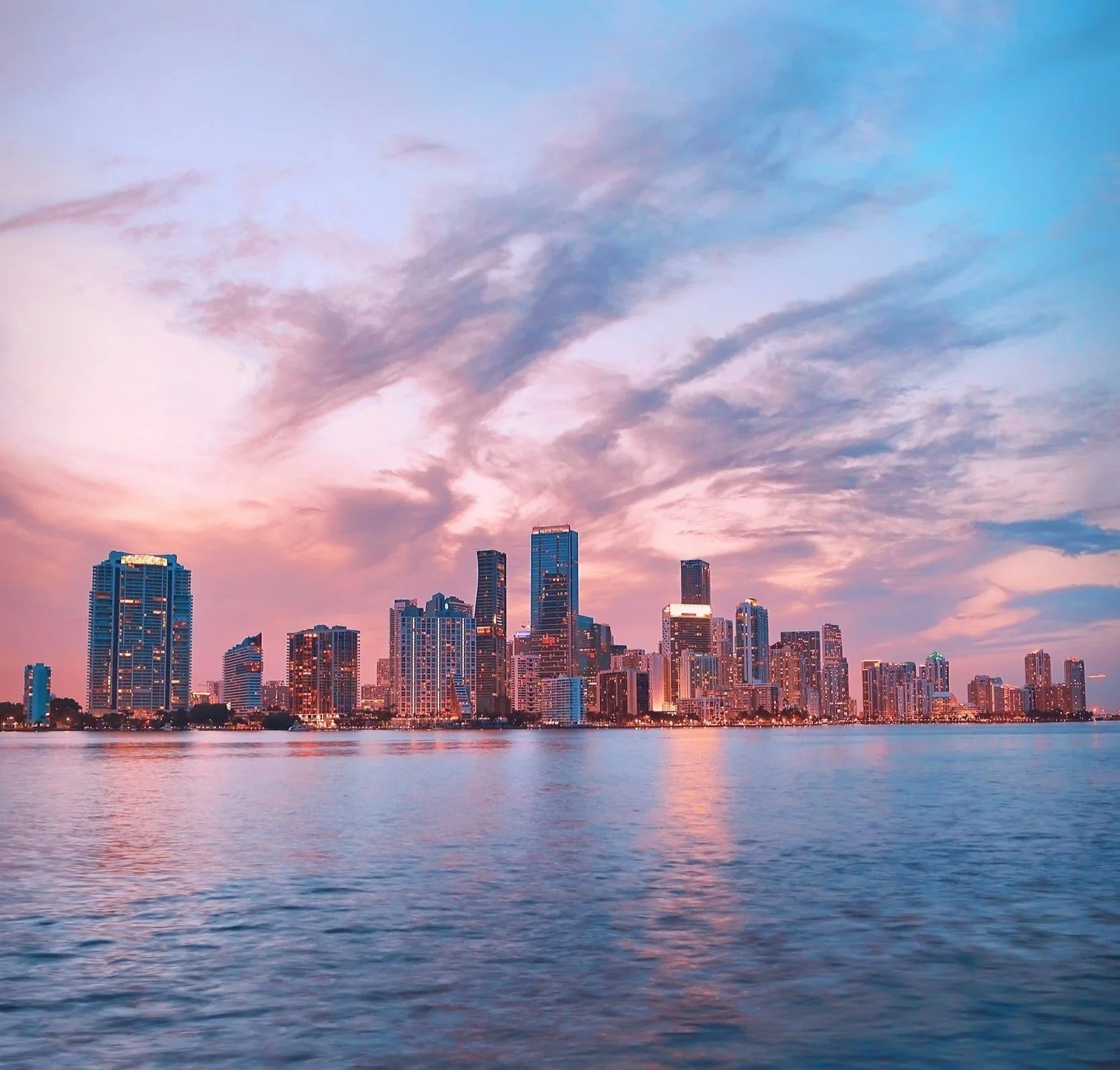 Skyline of a city with tall buildings at sunset, reflecting on a body of water in the foreground.