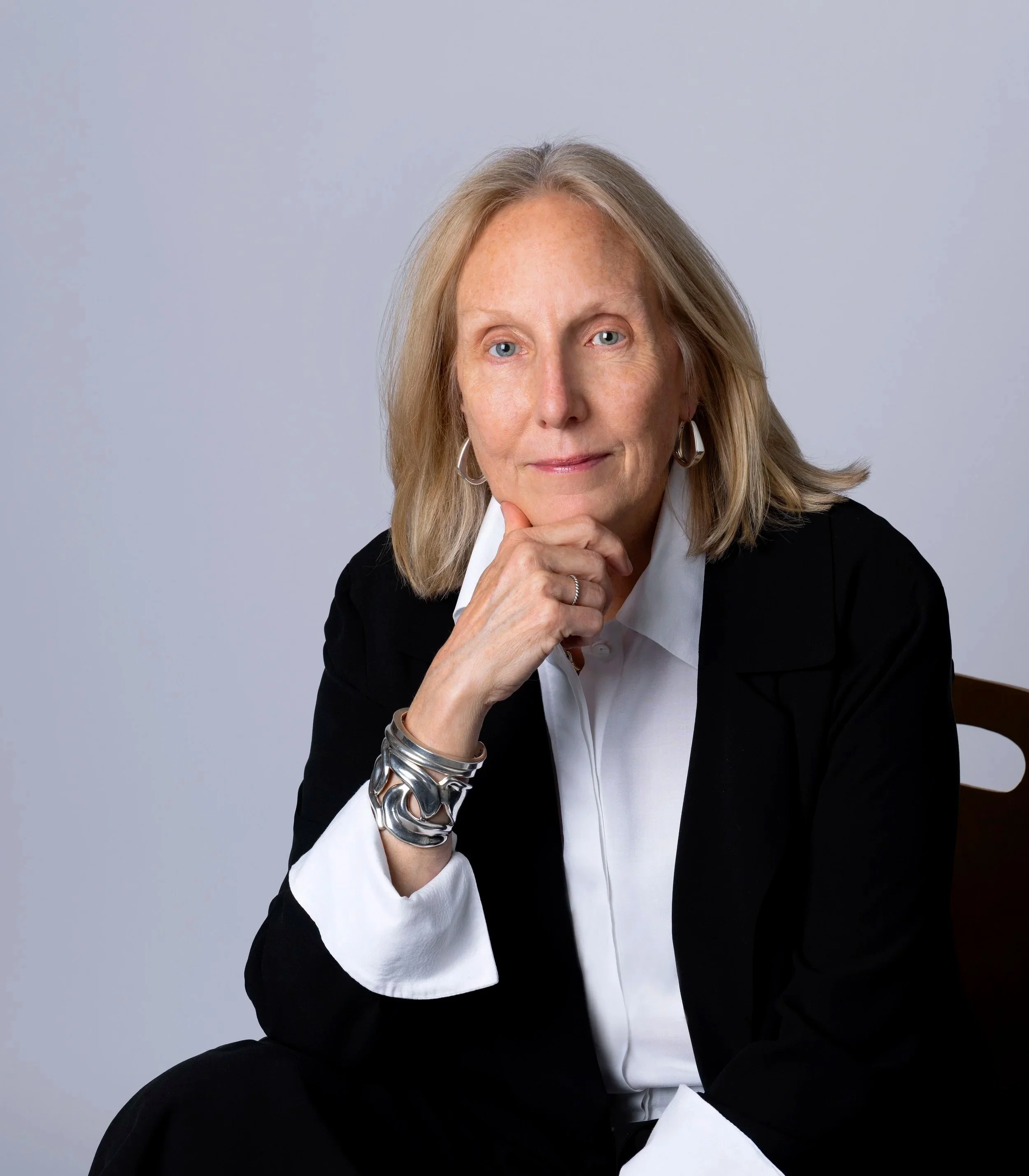 A middle-aged woman with blonde hair, wearing a black blazer and white shirt, sitting with her chin resting on her hand, looking at the camera with a confident expression.