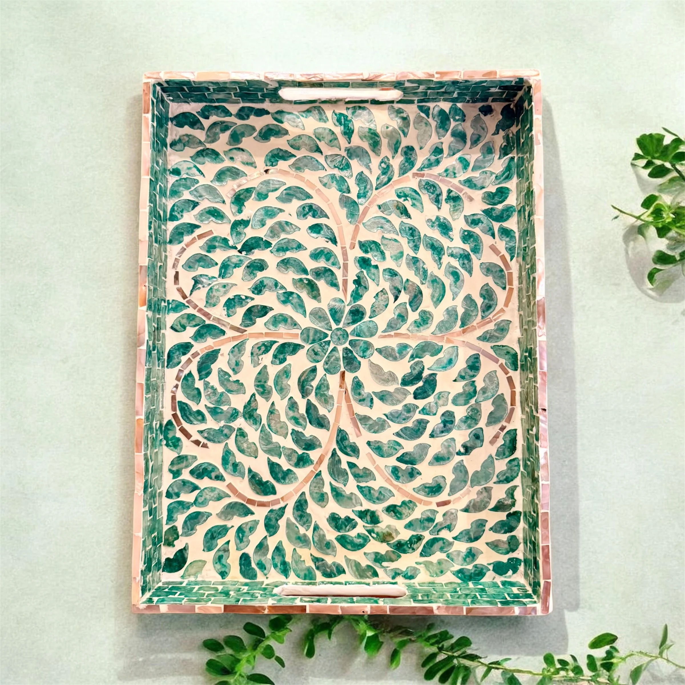 Decorative rectangular tray with a floral and leaf mosaic pattern in shades of green, pink, and white, placed on a light green surface with some small green plants around.