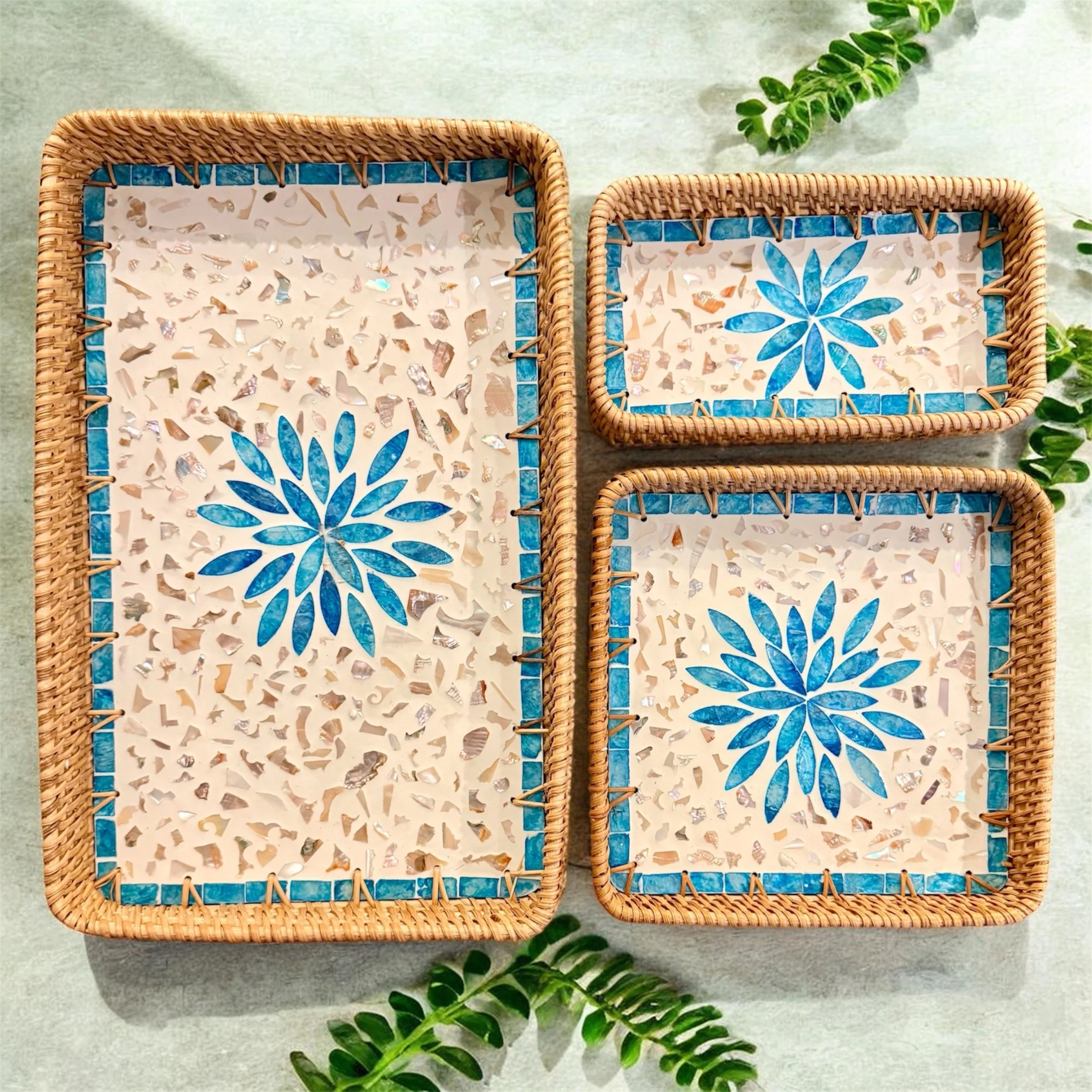 Three rectangular wicker trays with mosaic tile designs featuring blue floral patterns, placed on a light surface with green foliage nearby.