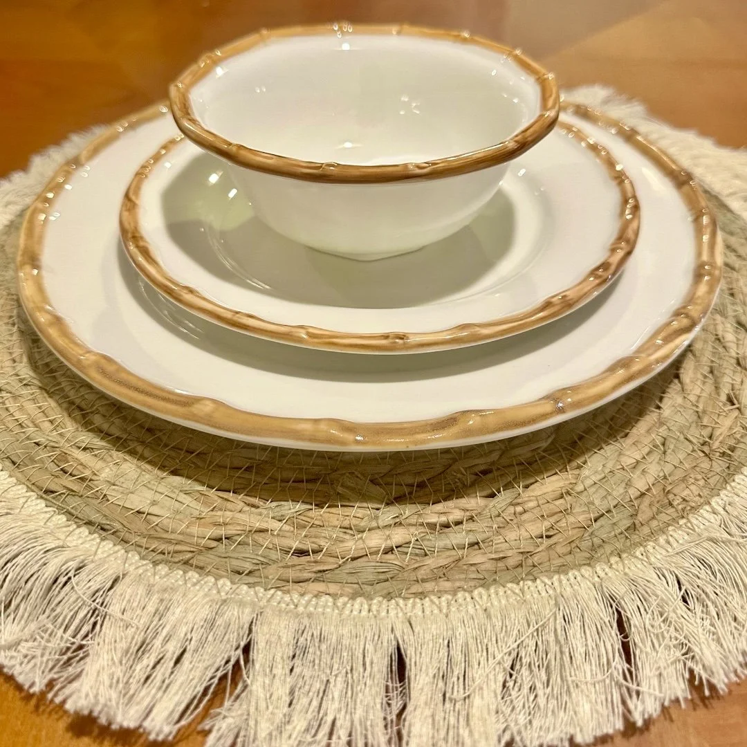 A white ceramic tea set with brown trim, consisting of a tea cup, saucer, and serving plate, placed on a woven placemat with fringes, on a wooden table.