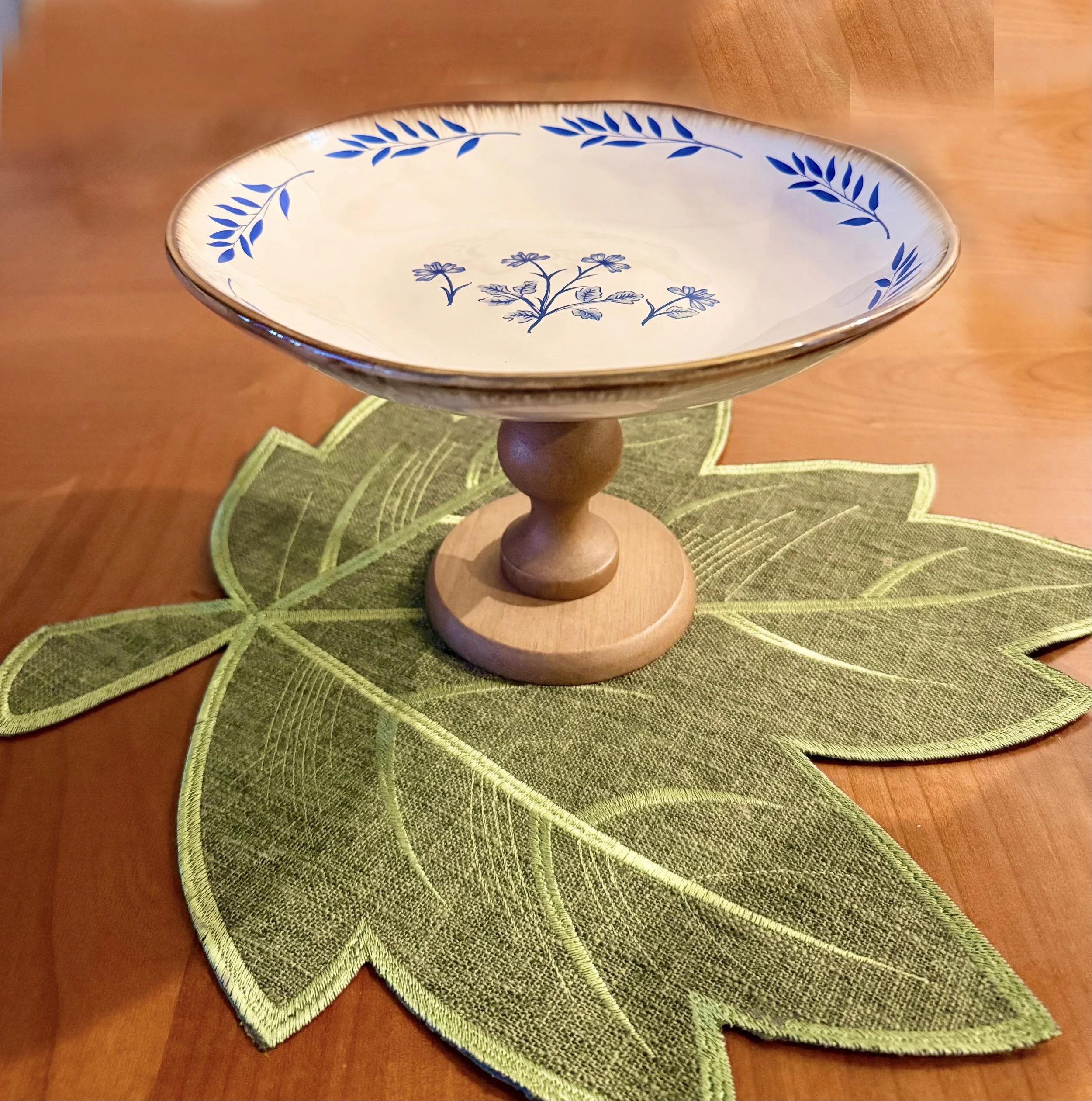 A ceramic cake stand with a blue and white floral pattern is placed on a green leaf-shaped placemat on a wooden table.