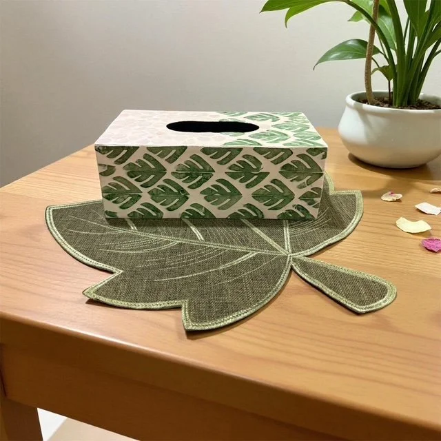 Tissue box with leaf pattern on a leaf-shaped placemat on a wooden table, with a potted plant and scattered flower petals in the background.