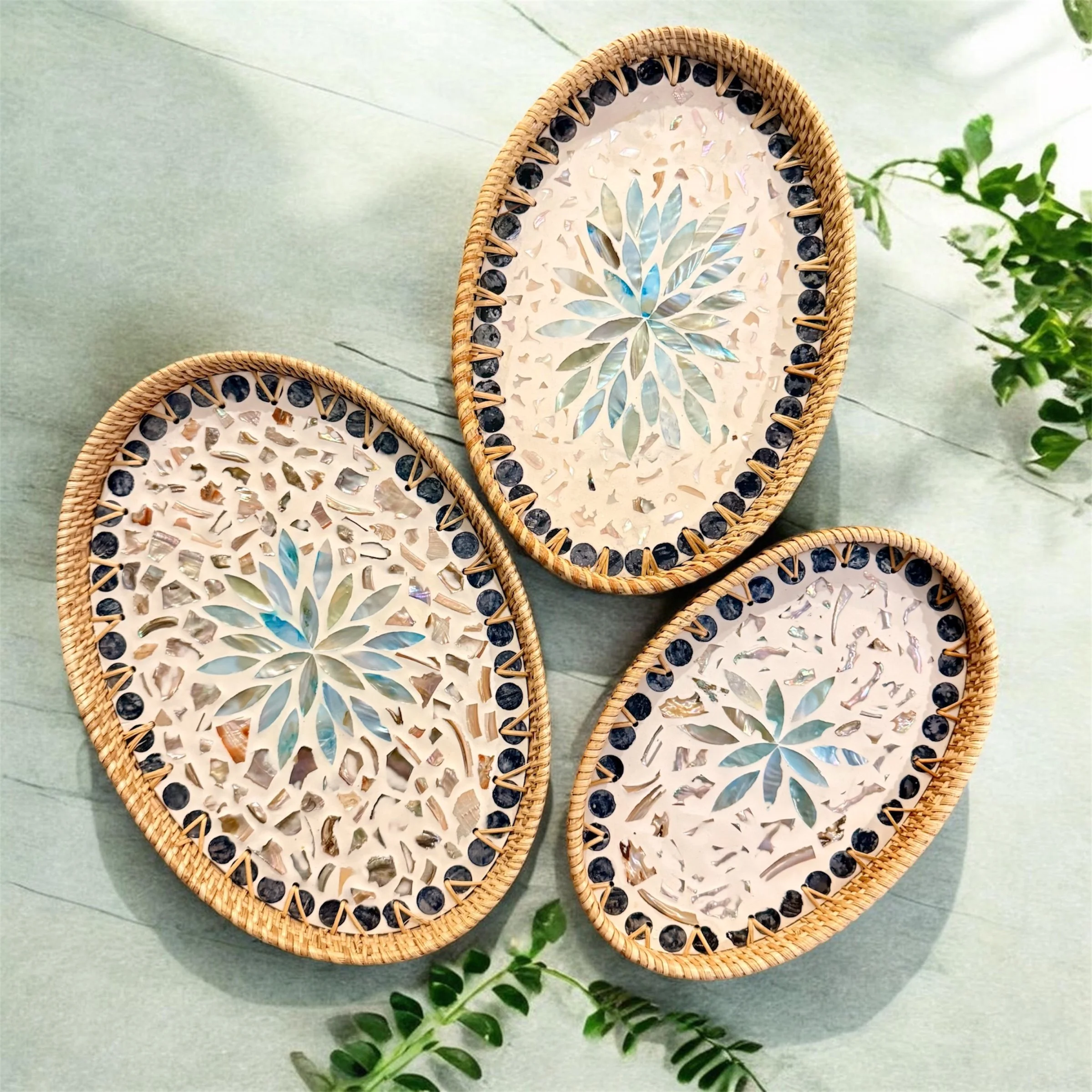 Three oval rattan trays with mosaic sea shell designs and black bead borders, placed on a light green surface with some green leaves.