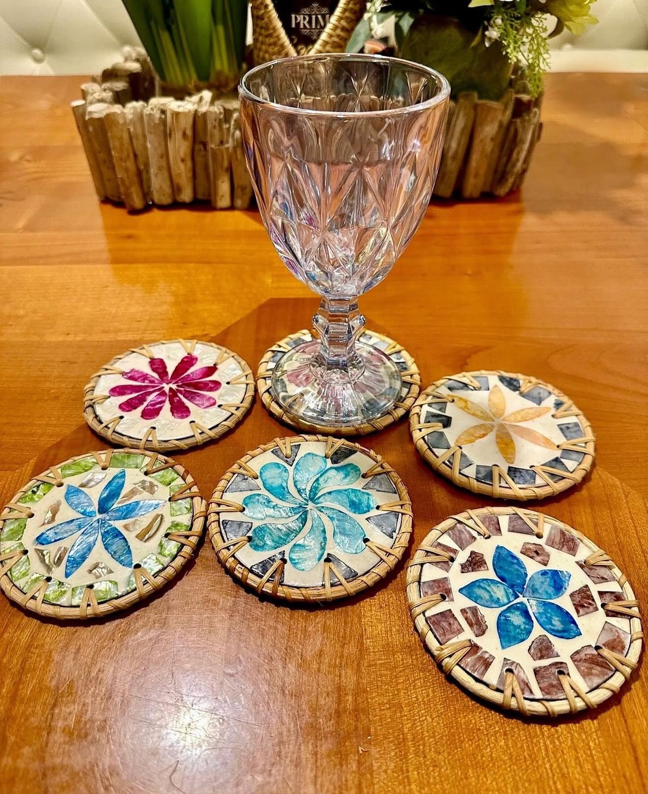 A clear, faceted glass on a wooden table, surrounded by five colorful mosaic coasters with floral patterns and woven edges, with a decorative plant in the background.