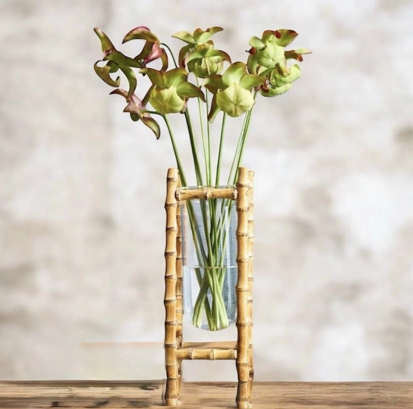 A glass vase filled with green and pinkish flowers, placed on a small bamboo stand on a wooden surface.