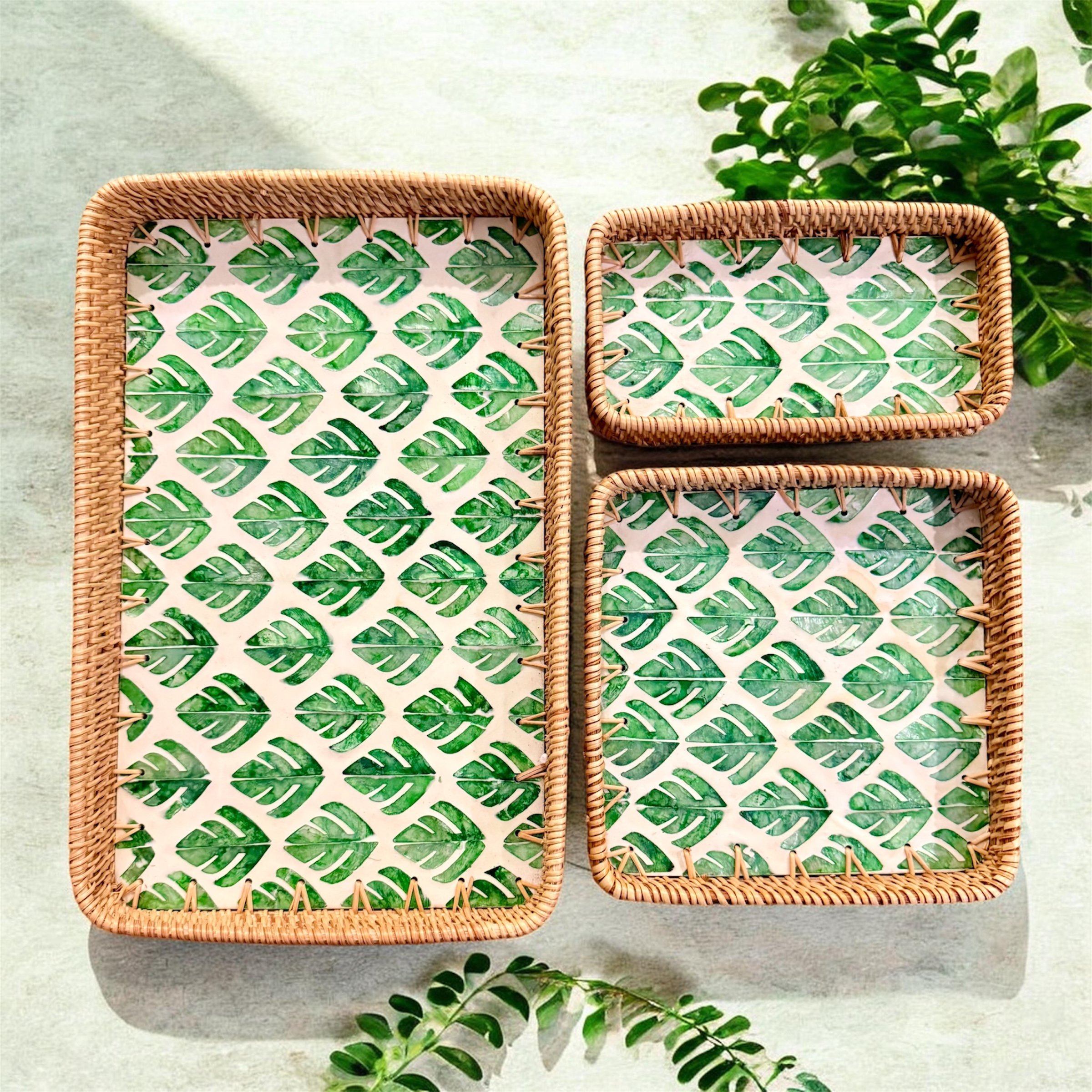Three rectangular woven rattan trays with leaf-patterned liners on a light surface, with green foliage nearby.